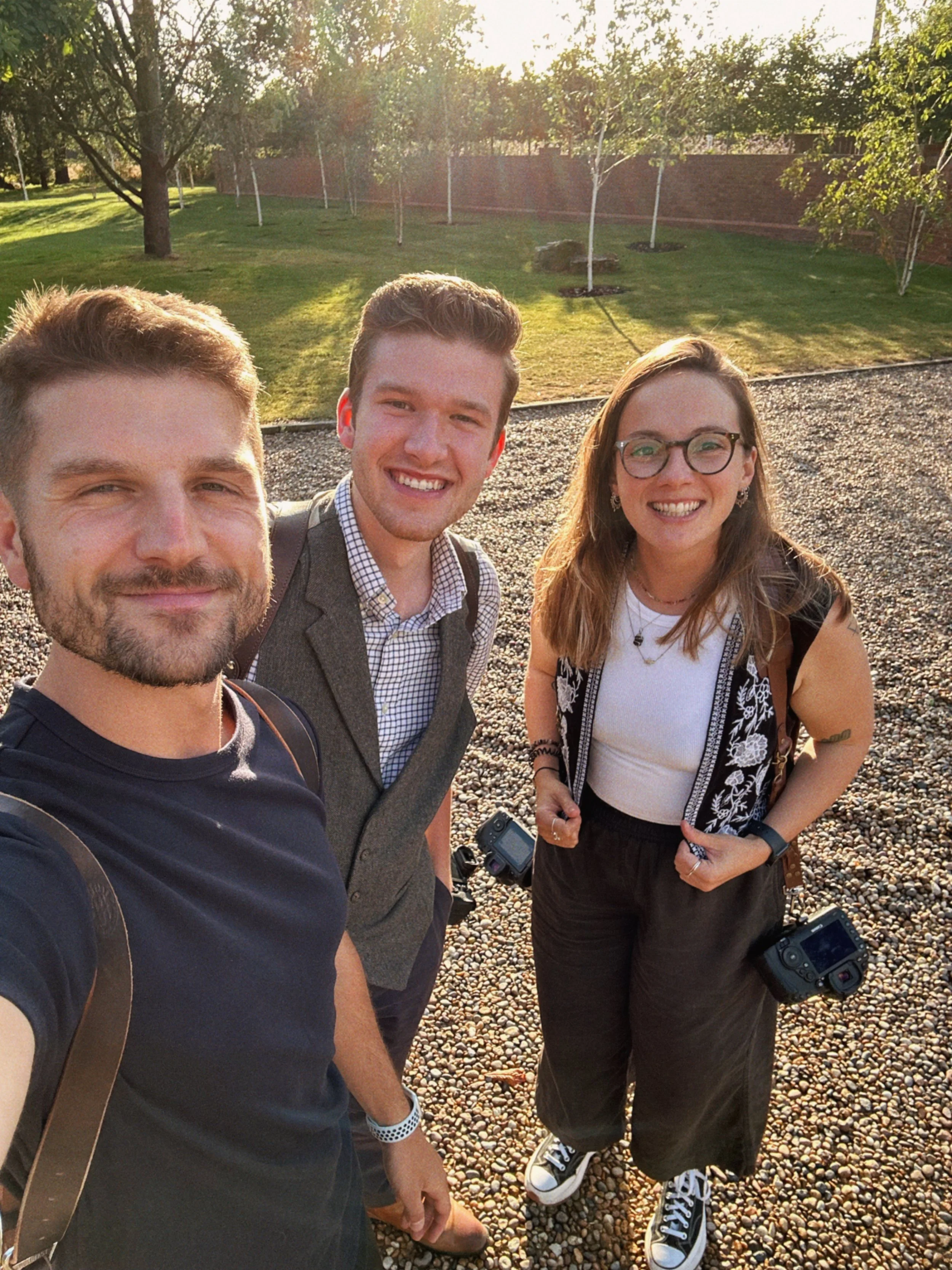 Three young adults smiling and posing for a selfie outdoors on a sunny day, with a grassy area, trees, and a brick fence in the background.