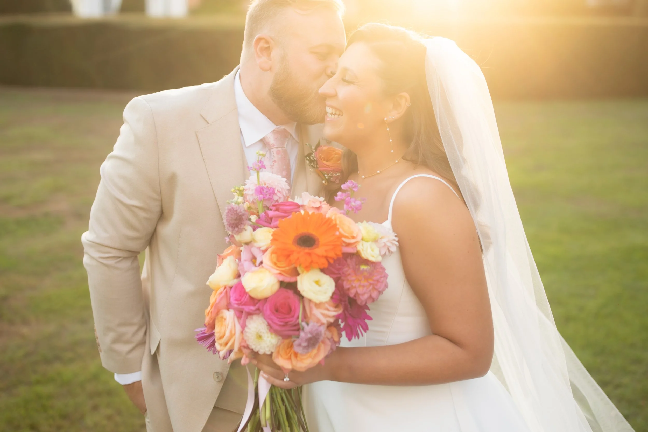 A bride and groom sharing a kiss on their wedding day, outdoors with sunlight shining in the background. The bride holds a vibrant bouquet of pink, orange, and white flowers, and wears a white wedding dress and veil. The groom wears a beige suit with a pink tie.