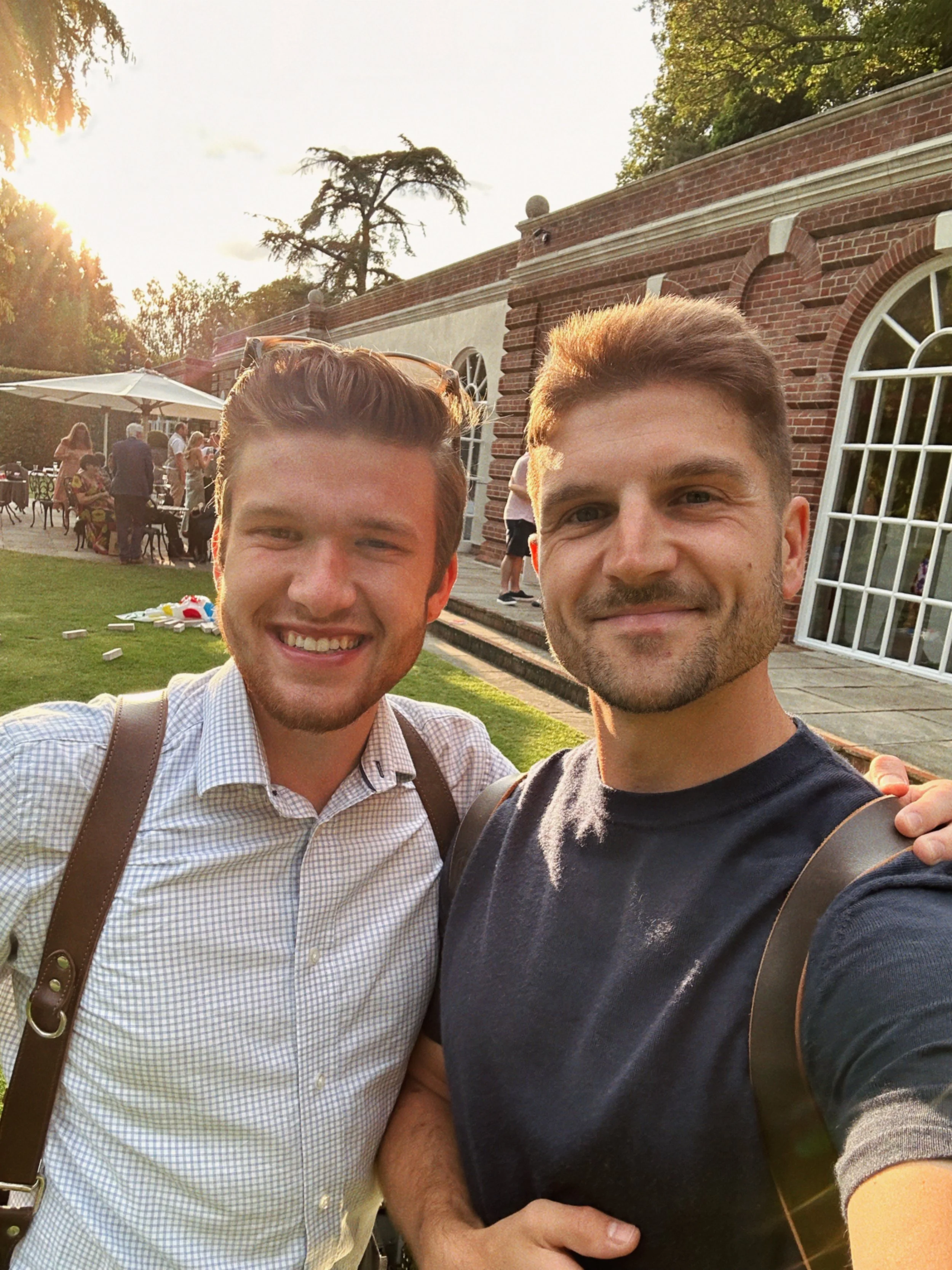 Two smiling men taking a selfie outdoors in a garden area with a brick building, people socializing, and tables with umbrellas in the background during golden hour.