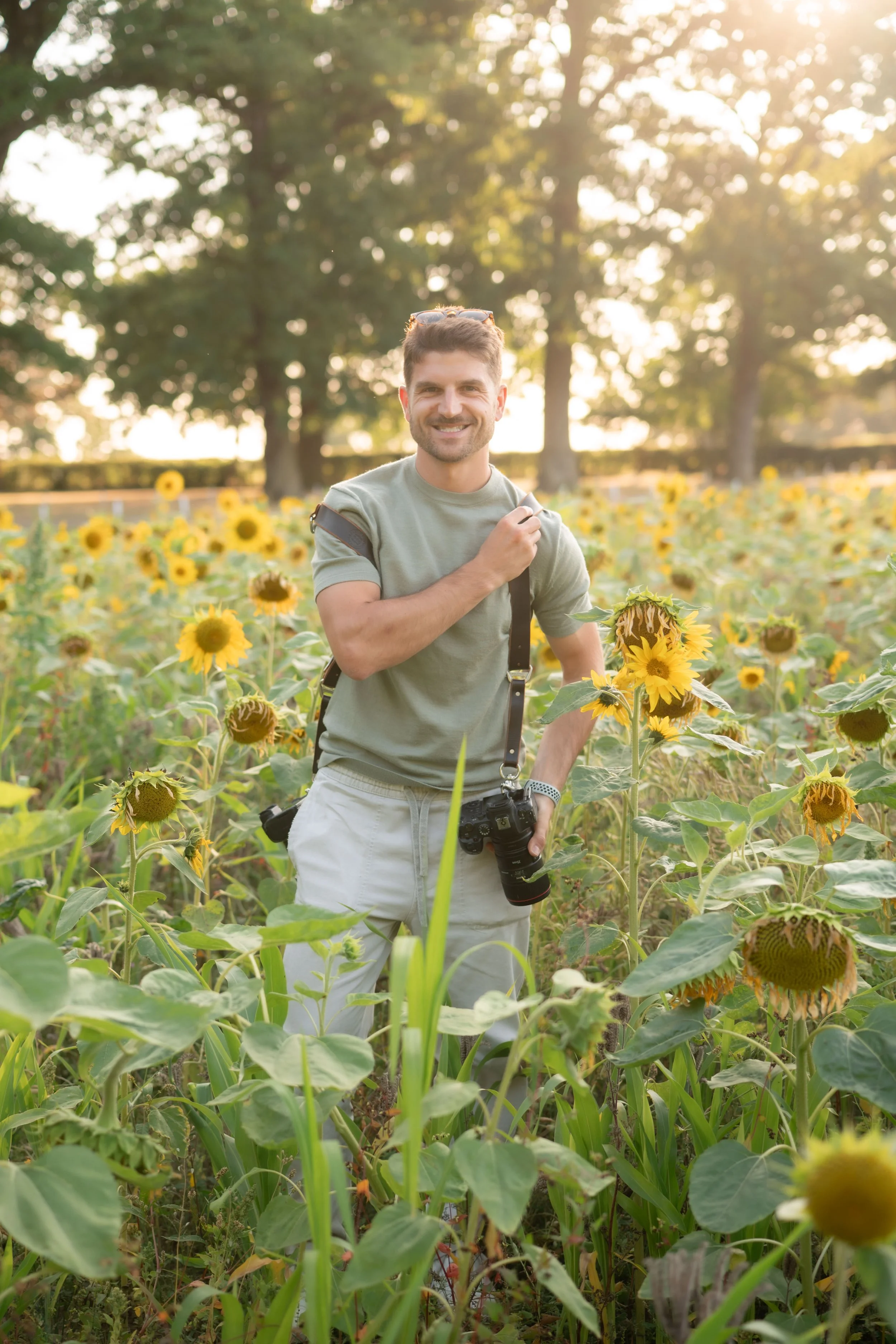 A man standing in a sunflower field during sunset, smiling, wearing a green t-shirt and white shorts, with a camera hanging around his neck.