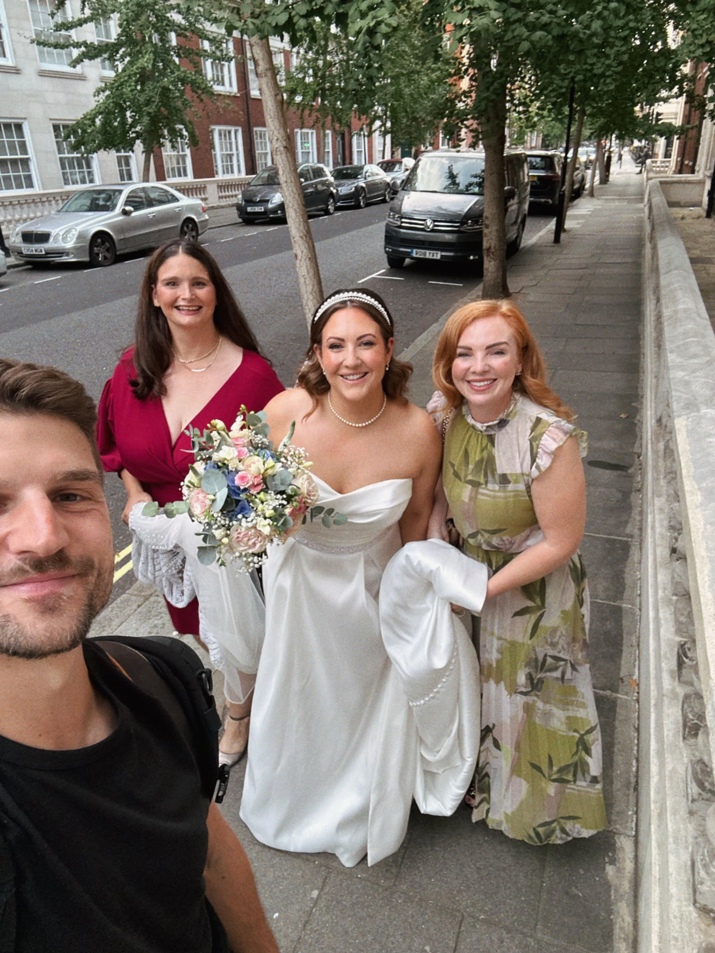 Four people, including a bride in a white wedding dress holding a bouquet, are standing on a sidewalk in an urban area with parked cars and trees in the background. They are smiling and taking a selfie.