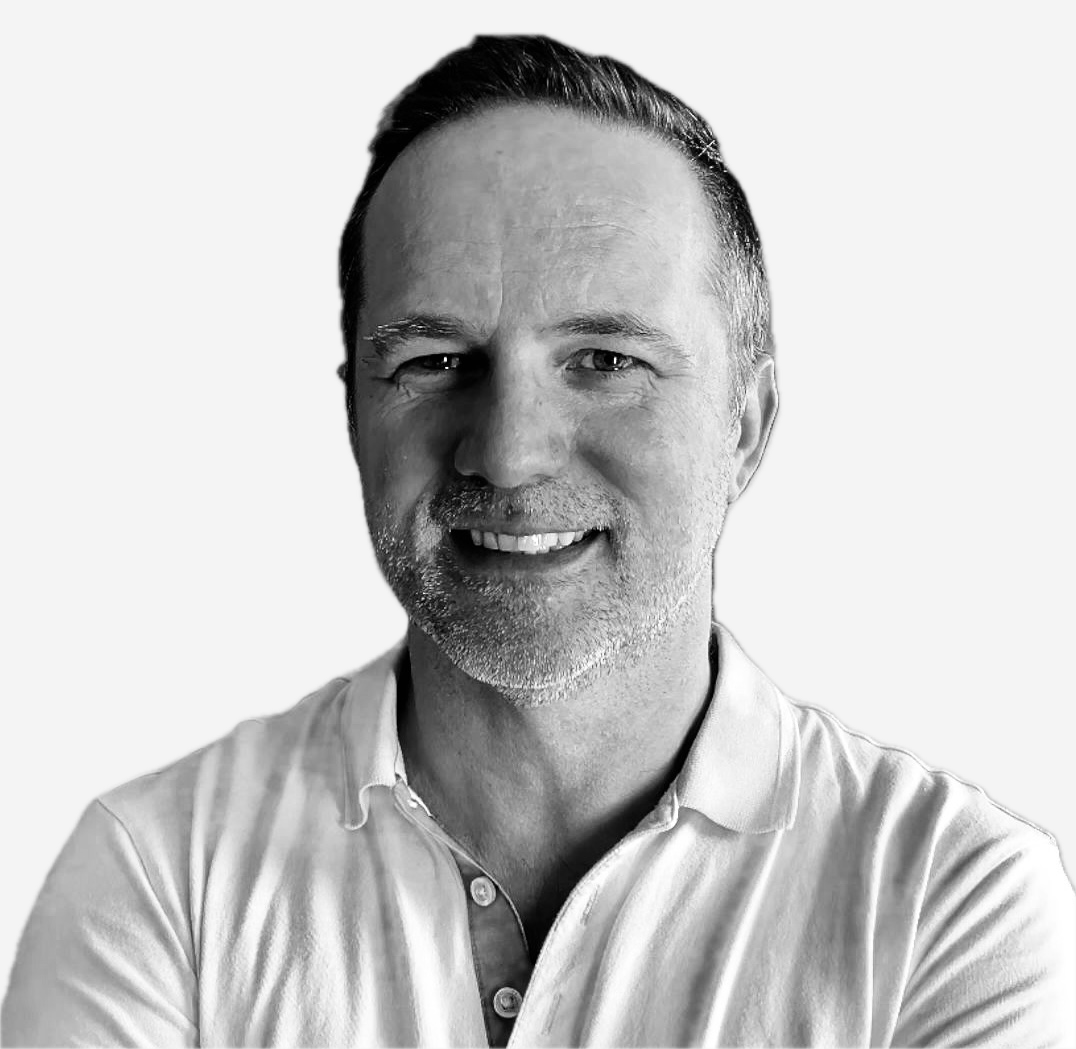 Black and white photo of a smiling man with short hair, wearing a light-colored collared shirt, against a plain light background. Scott Stonham