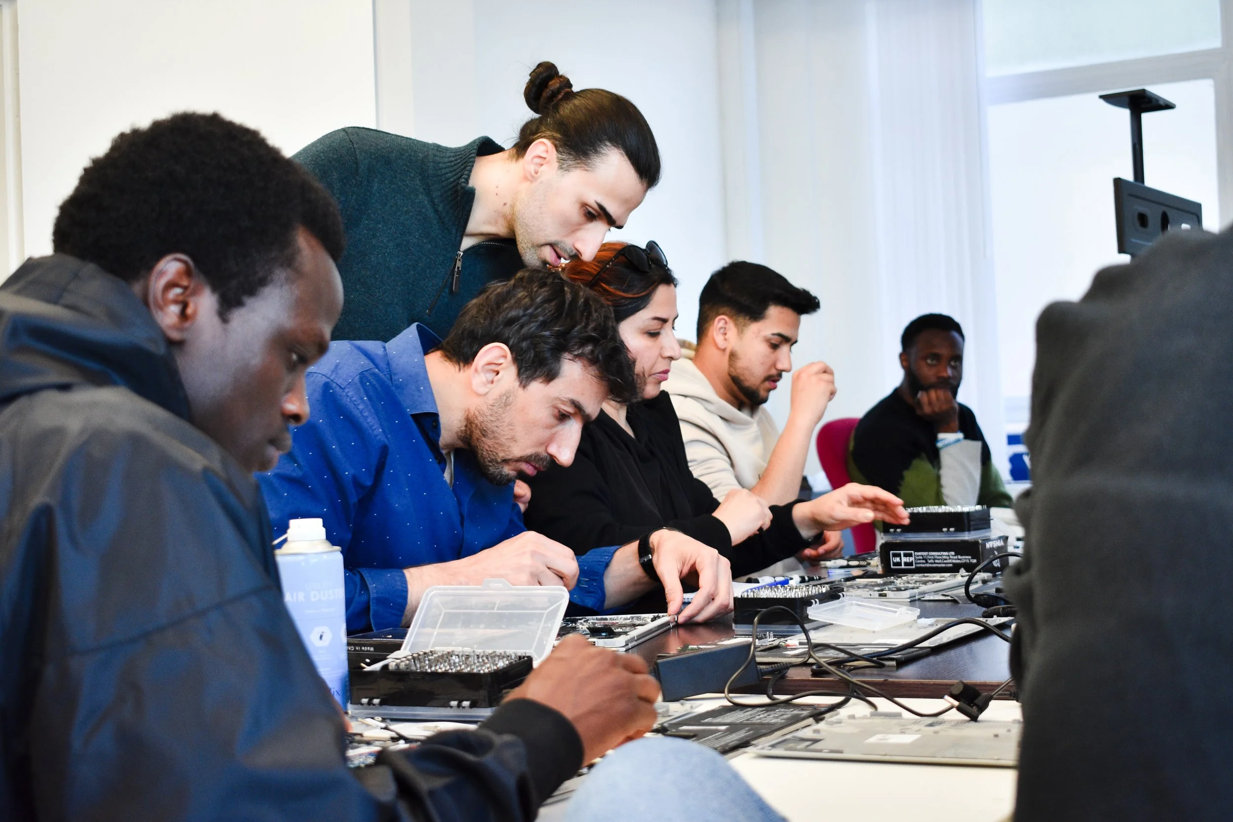 Group of six young adults working on electronics repair or assembly at a table, with tools and parts, in a well-lit room.