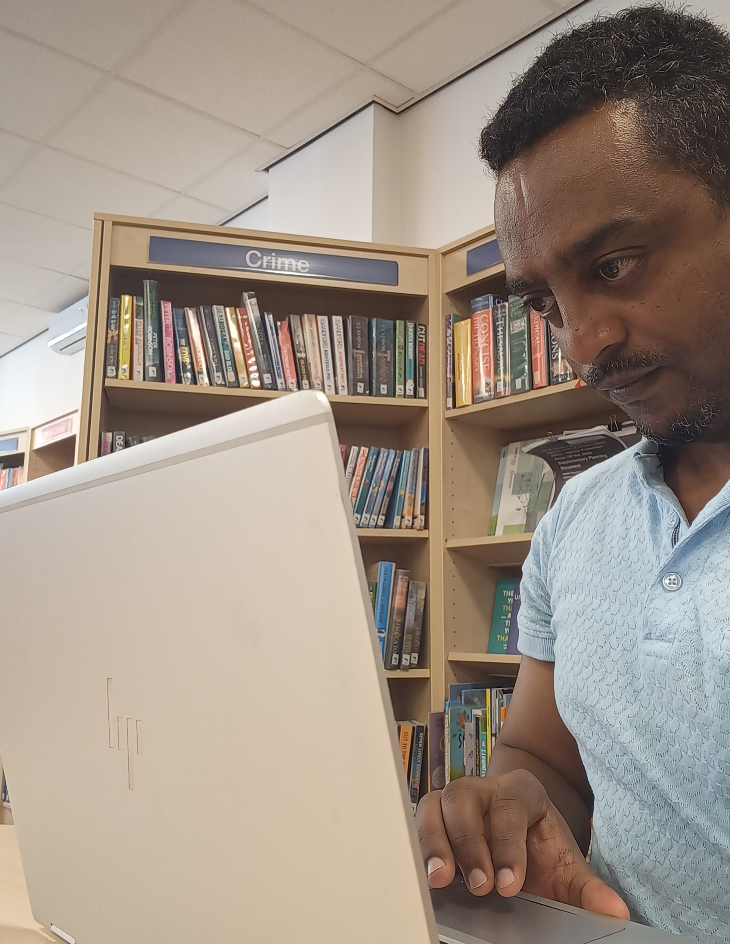 A man in a light blue shirt working hard, using a laptop in a library.