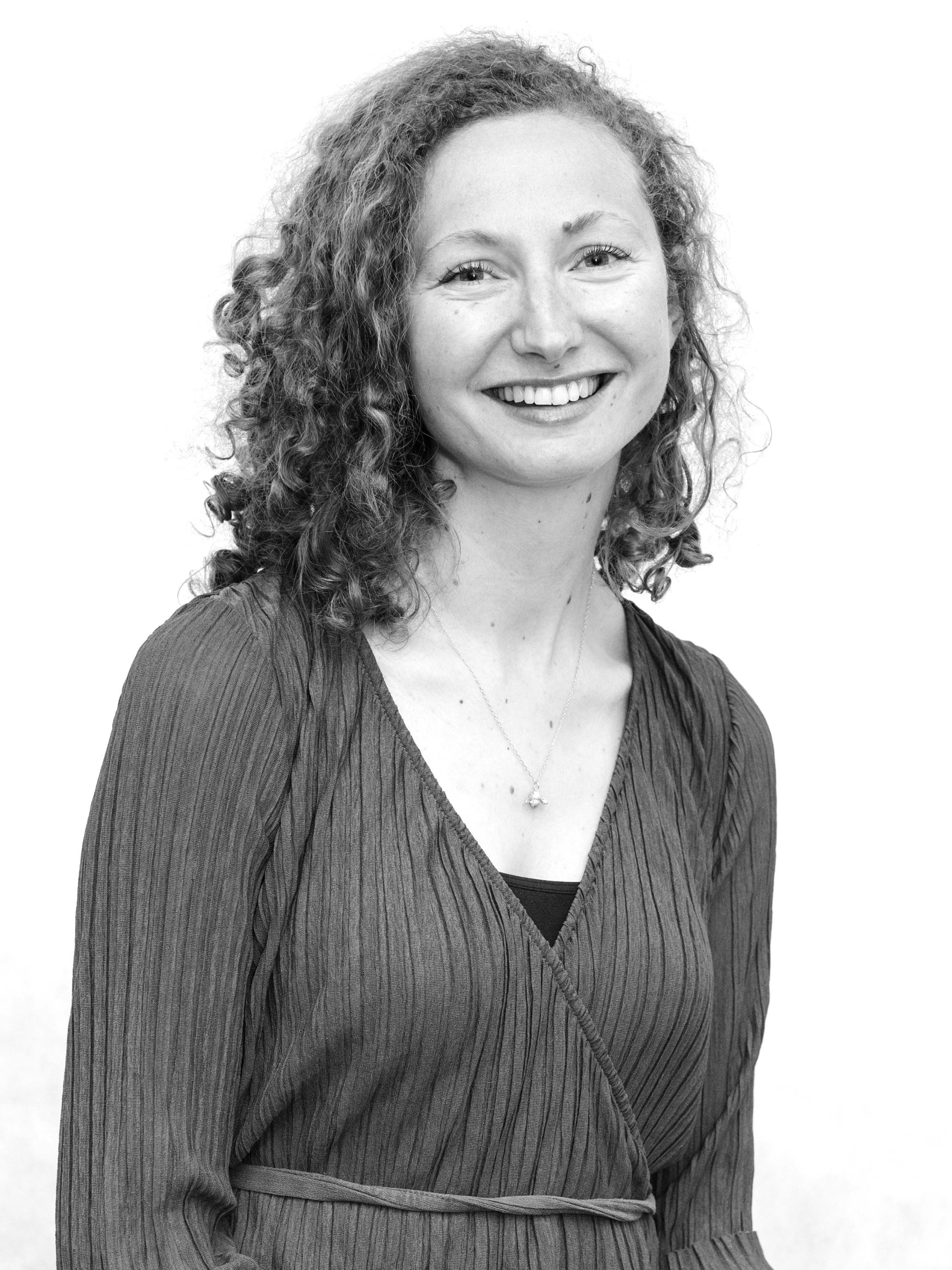 Black and white portrait of a smiling woman with curly hair, wearing a textured dress with a V-neck and a necklace. Suzie Hance-Barkley