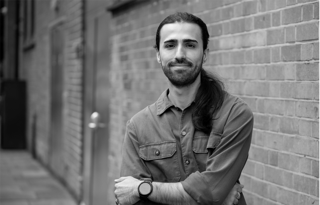 Black and white photo of a man with long hair, beard, and mustache, standing with his arms crossed, smiling, next to a brick wall. Parviz Sedighi