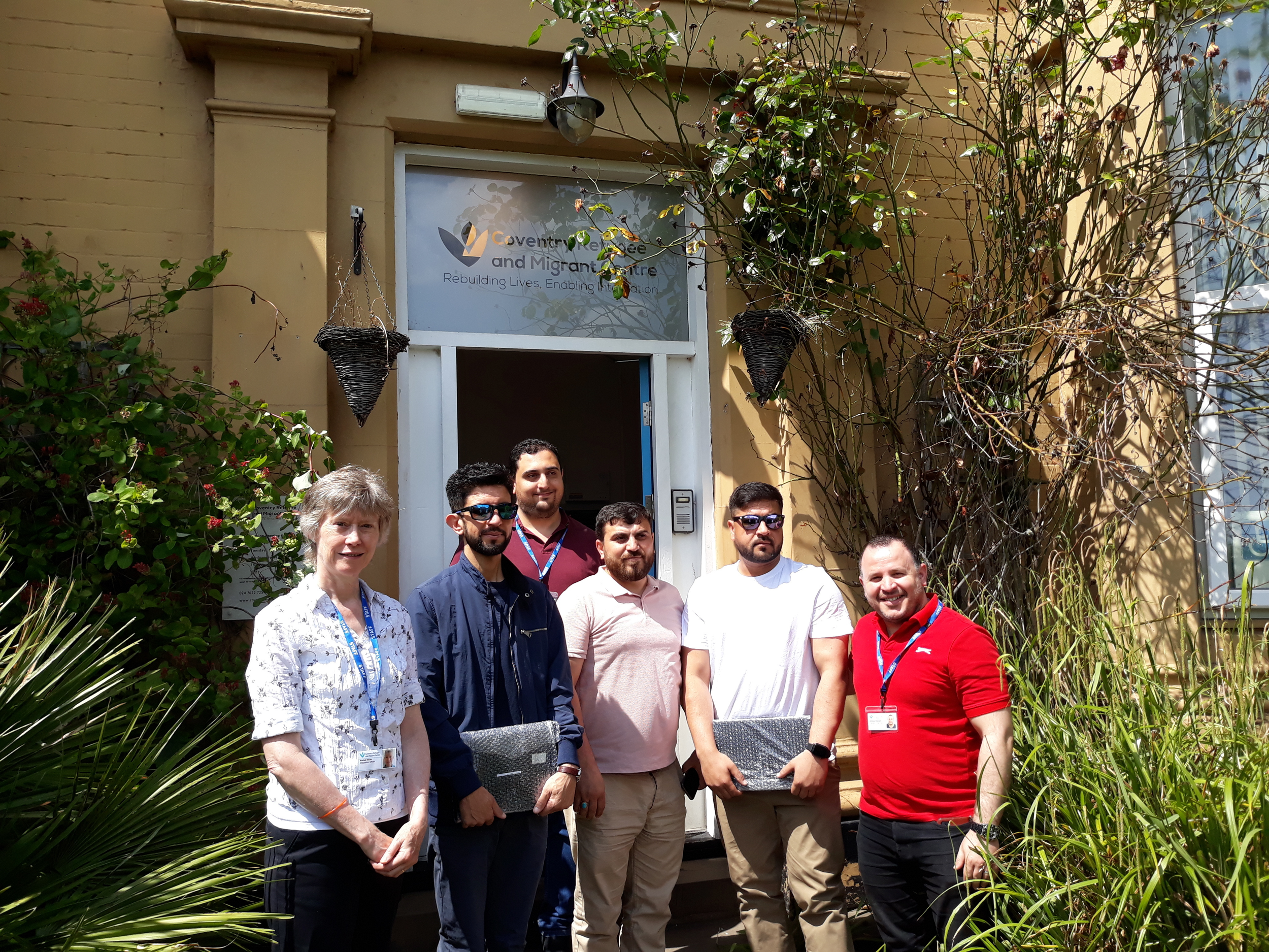 Group of six people standing outside Coventry Refugee and Migrant Centre, in front of the entrance with lush greenery on both sides.
