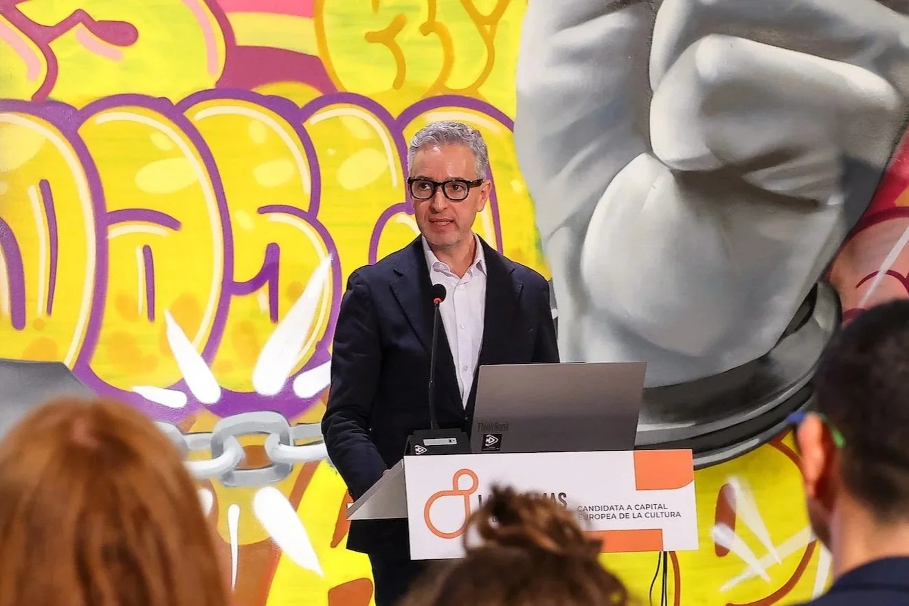 José Luis Pérez Pont habla desde un atril con un ordenador portátil, ante un mural de grafiti con un puño de piedra y cadenas