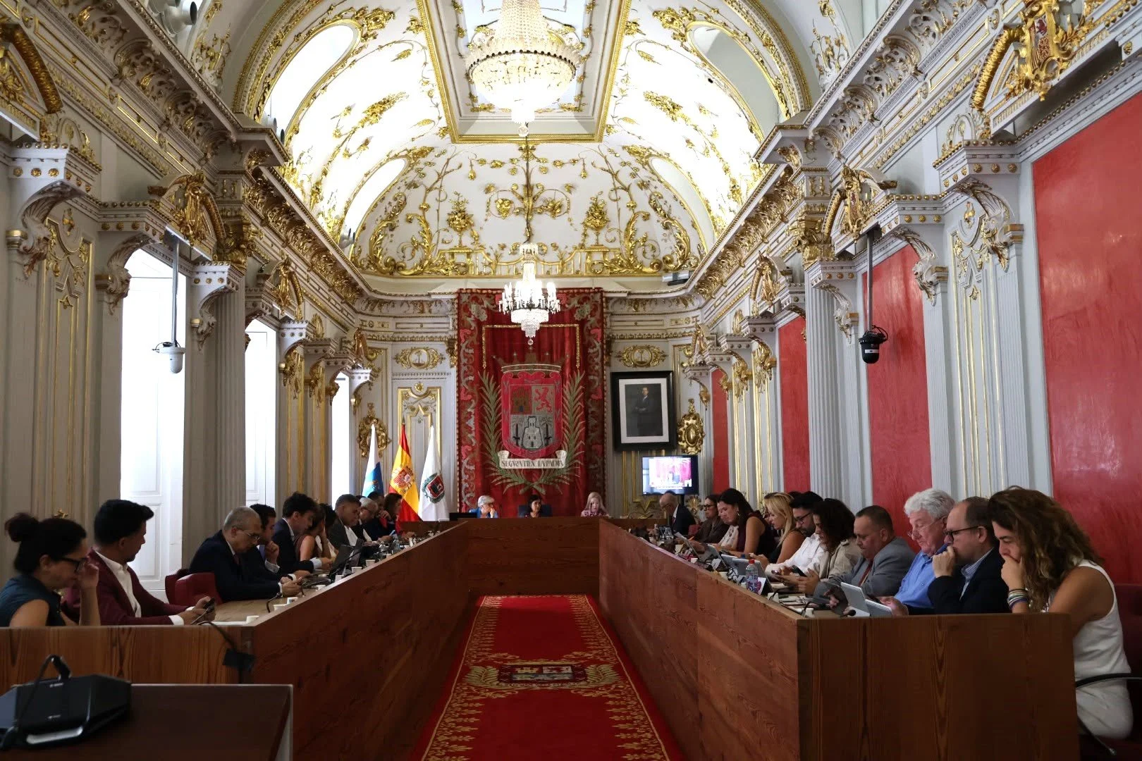 Sesión plenaria en el salón de plenos de las Casas Consistoriales, de techos dorados y paredes rojas, con los concejales sentados a ambos lados de una larga mesa de madera en forma de U y la presidencia al fondo, bajo el estandarte de la ciudad