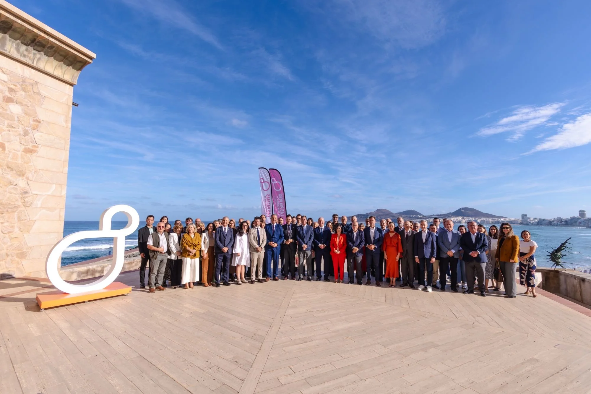 Foto de grupo de decenas de personas en la terraza del Auditorio Alfredo Kraus, junto al mar, con la escultura blanca del símbolo de LPGC’31 a la izquierda y la playa de Las Canteras al fondo