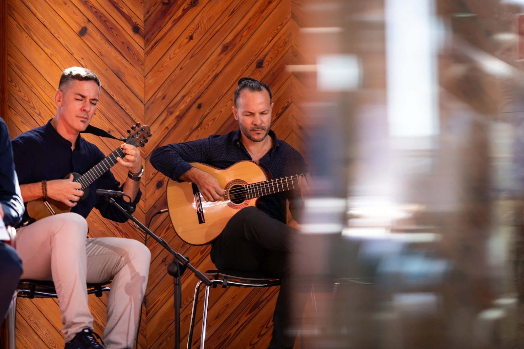 Dos guitarristas tocan sentados sobre un escenario, con una pared de madera en zigzag al fondo, durante una de las actuaciones showcase de WOMEX Gran Canaria