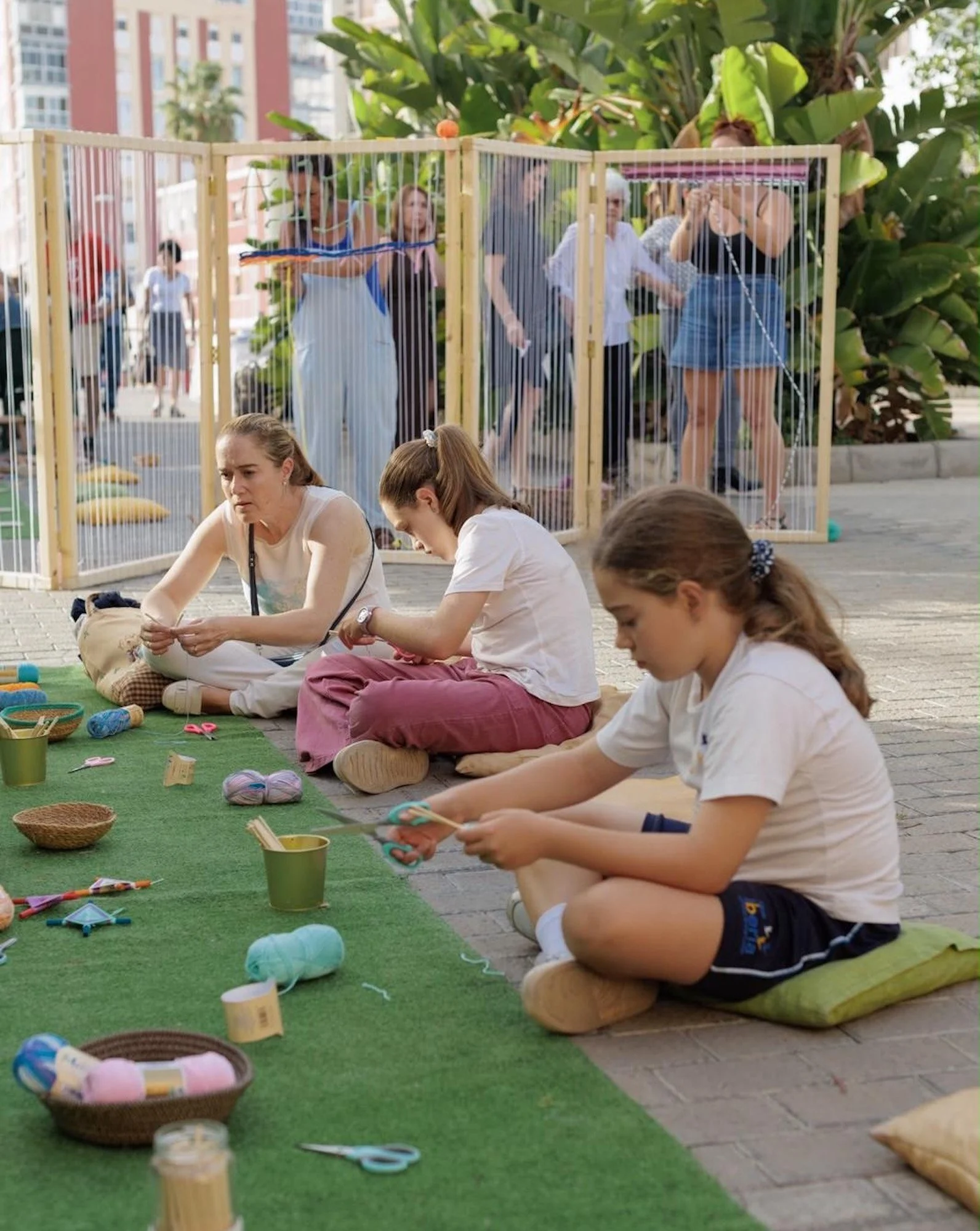 Una mujer y dos niñas tejen con hilos de colores en telares de madera durante una actividad familiar en la plaza