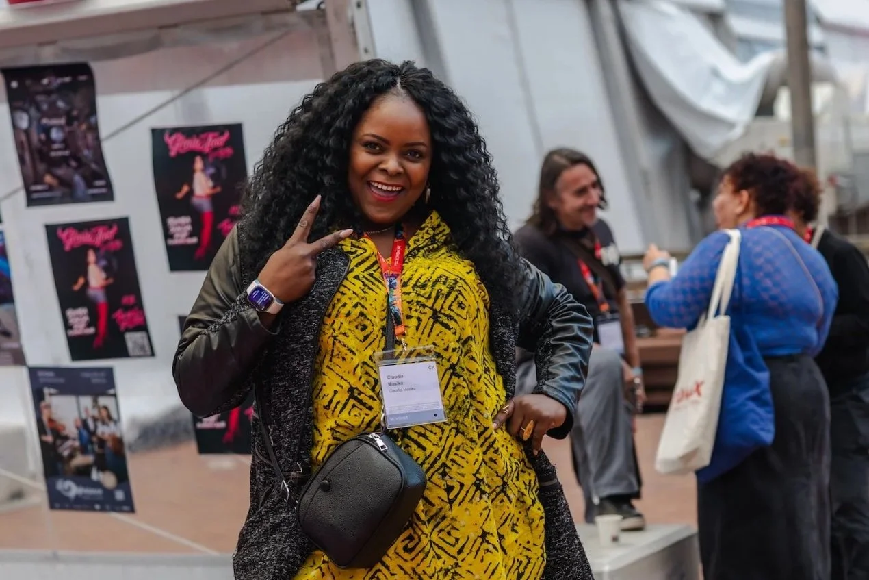 Una mujer con vestido amarillo estampado y acreditación de WOMEX sonríe a cámara haciendo el signo de la victoria, con otros asistentes y carteles de conciertos al fondo