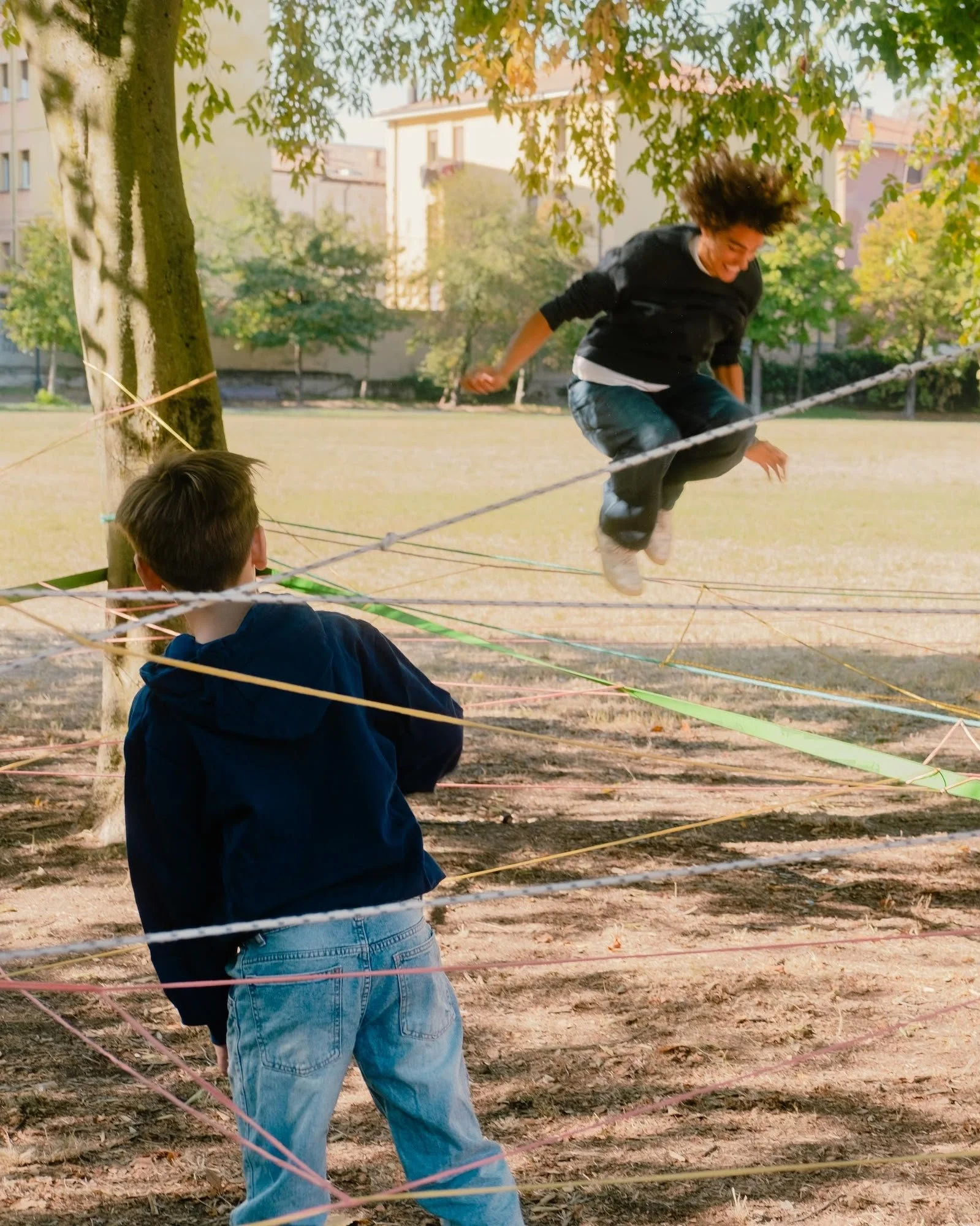 Una persona salta en el aire entre hilos de colores tensados entre los árboles de un parque, mientras otra la observa de espaldas