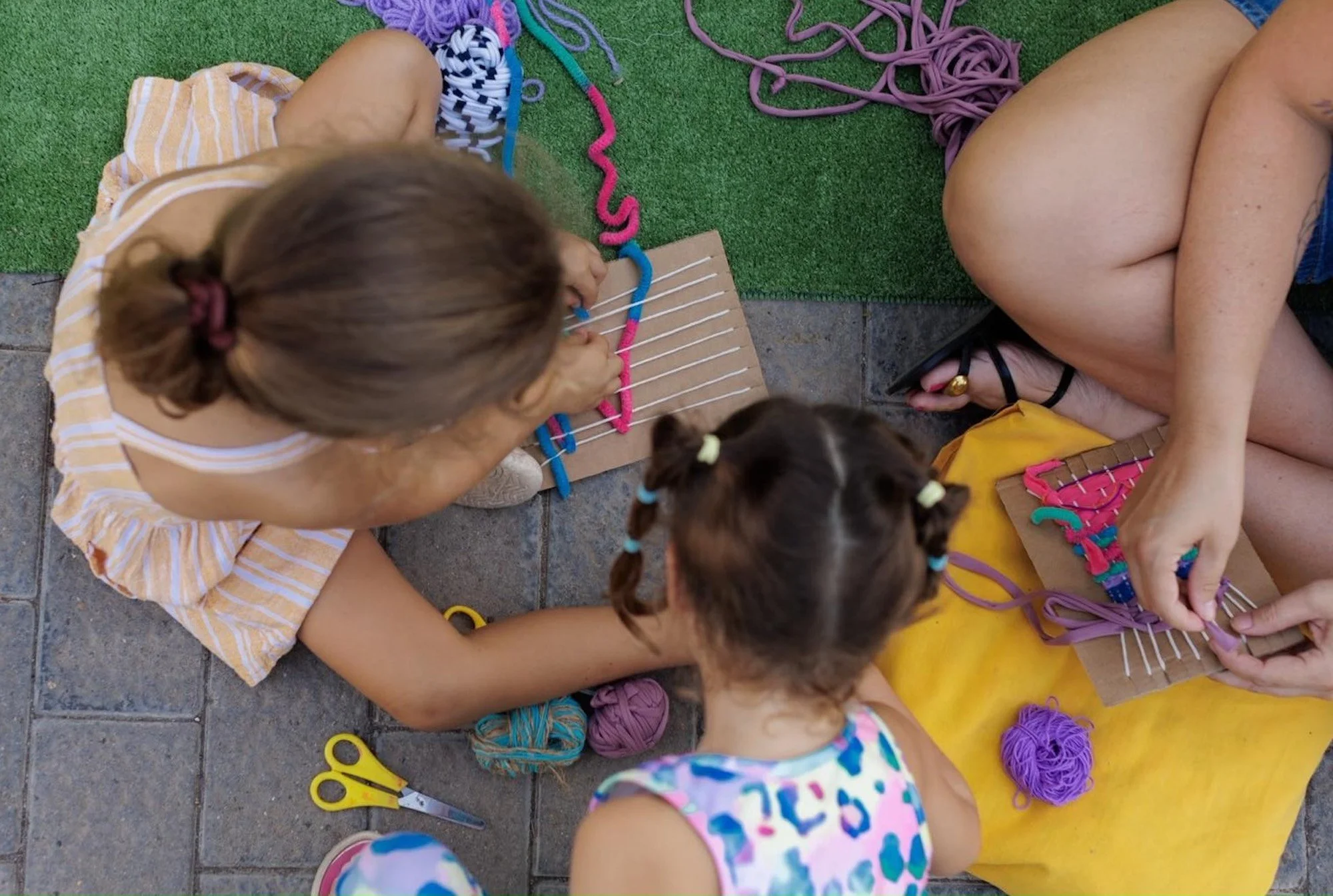 Dos niñas tejen con hilos de colores sobre telares de cartón caseros, vistas desde arriba