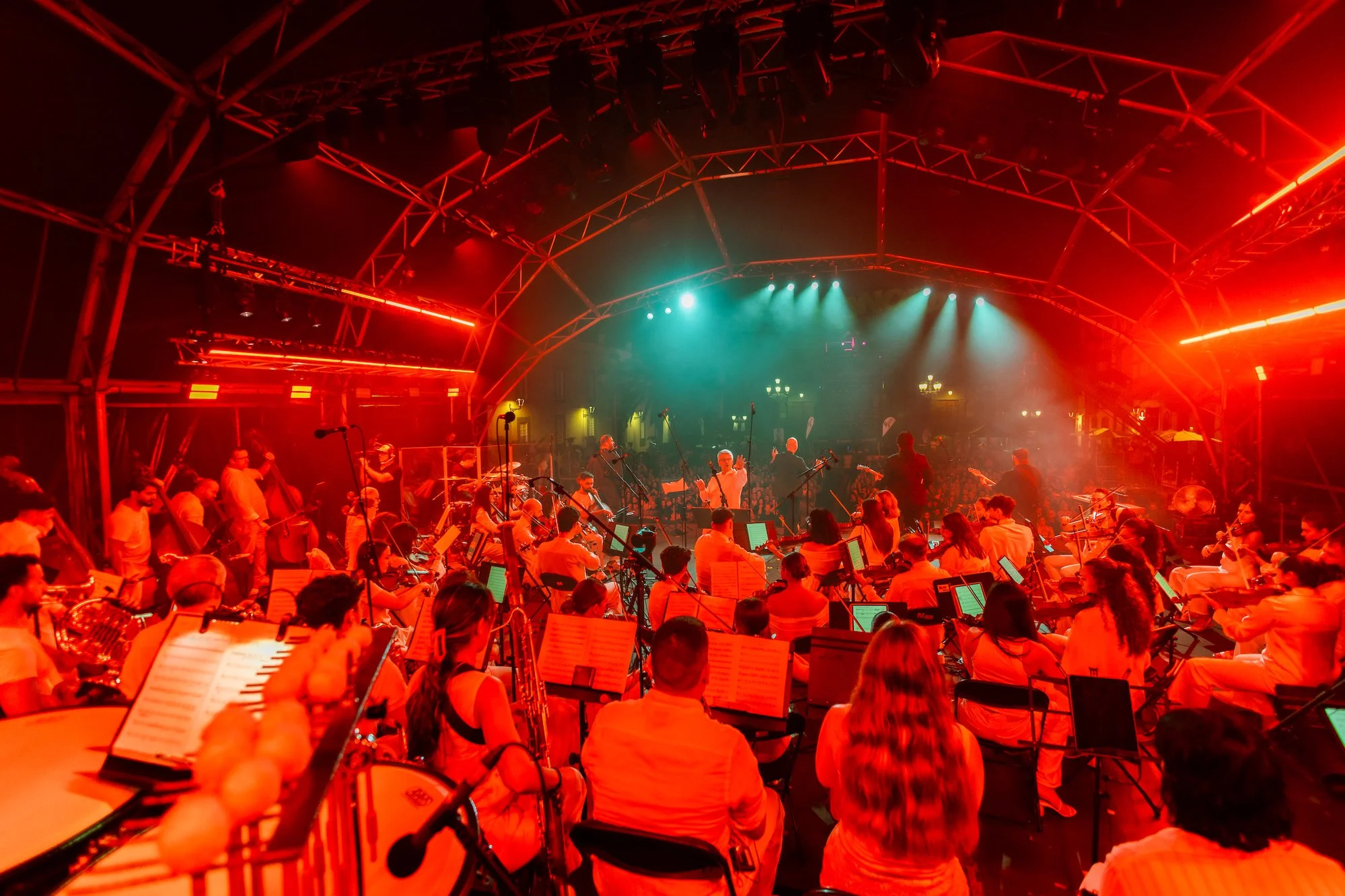 Una orquesta numerosa, vista desde detrás de los músicos, toca sobre un escenario cubierto e iluminado en rojo, con focos verdes al fondo