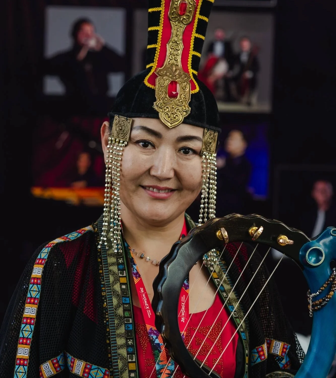 Una música sonríe a cámara vestida con un traje tradicional bordado y un alto tocado negro y rojo con adornos dorados y cadenas de cuentas, sosteniendo un instrumento de cuerda con forma de arpa