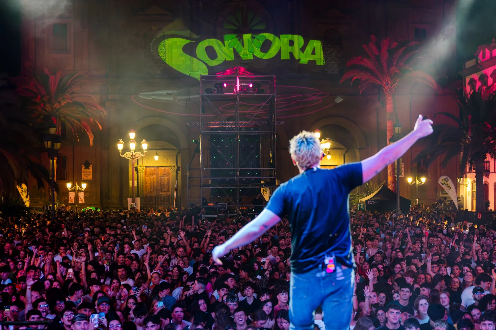 Un artista, de espaldas sobre el escenario, abre los brazos ante el público que llena la Plaza de Santa Ana de noche, con el logotipo de Sonora proyectado en verde sobre la fachada de la catedral