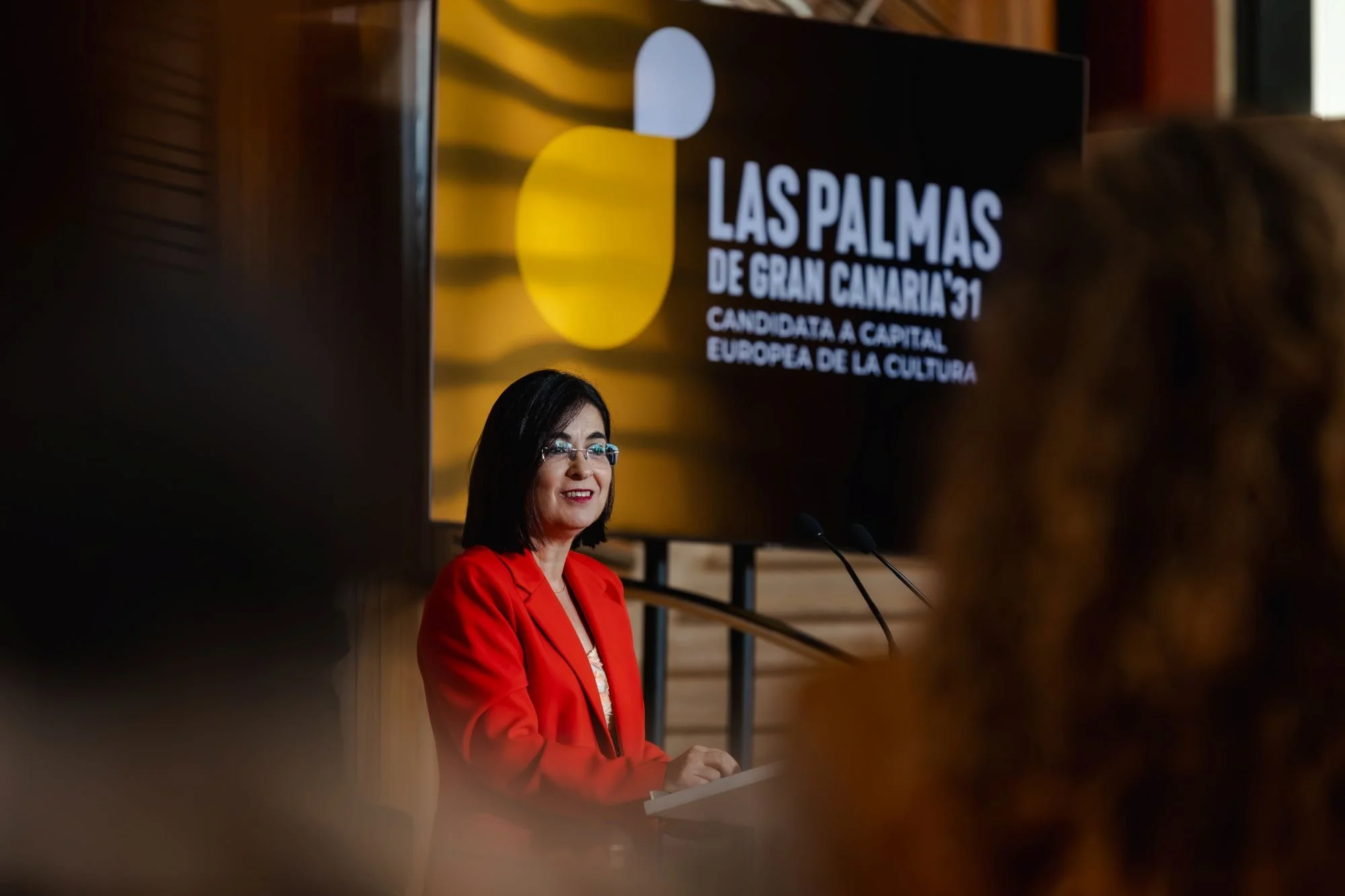 Carolina Darias interviene sonriente desde un atril, ante una pantalla con el logotipo de Las Palmas de Gran Canaria ‘31, con asistentes desenfocados en primer plano