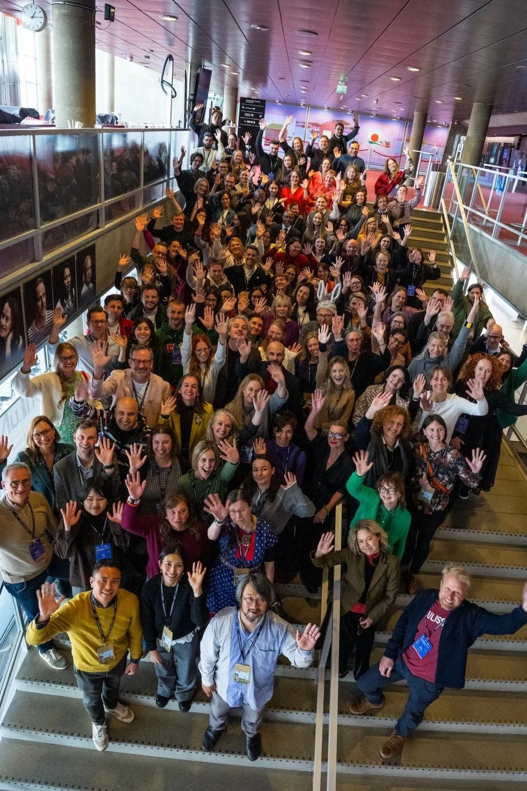 Foto de grupo tomada desde arriba de numerosos asistentes en una escalera interior, que saludan con la mano alzada mirando a la cámara