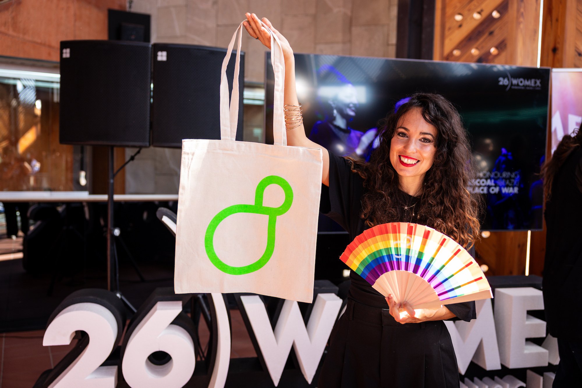 Una mujer sonríe junto a las letras gigantes '26 WOMEX', sosteniendo en alto una bolsa de tela con el logo del festival y un abanico de colores arcoíris