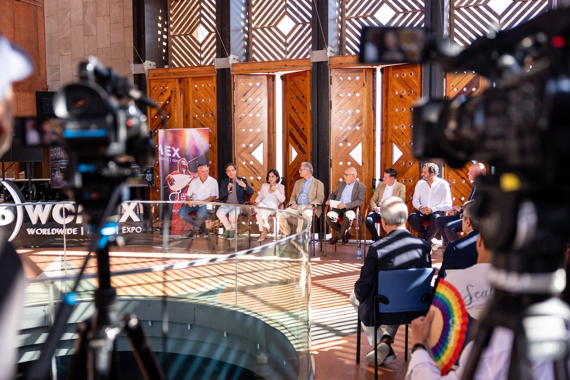 Varias personas conversan sentadas en un panel durante la presentación de WOMEX Gran Canaria, con cámaras de televisión grabando en primer plano y un cartel de 'WOMEX Expo' al fondo