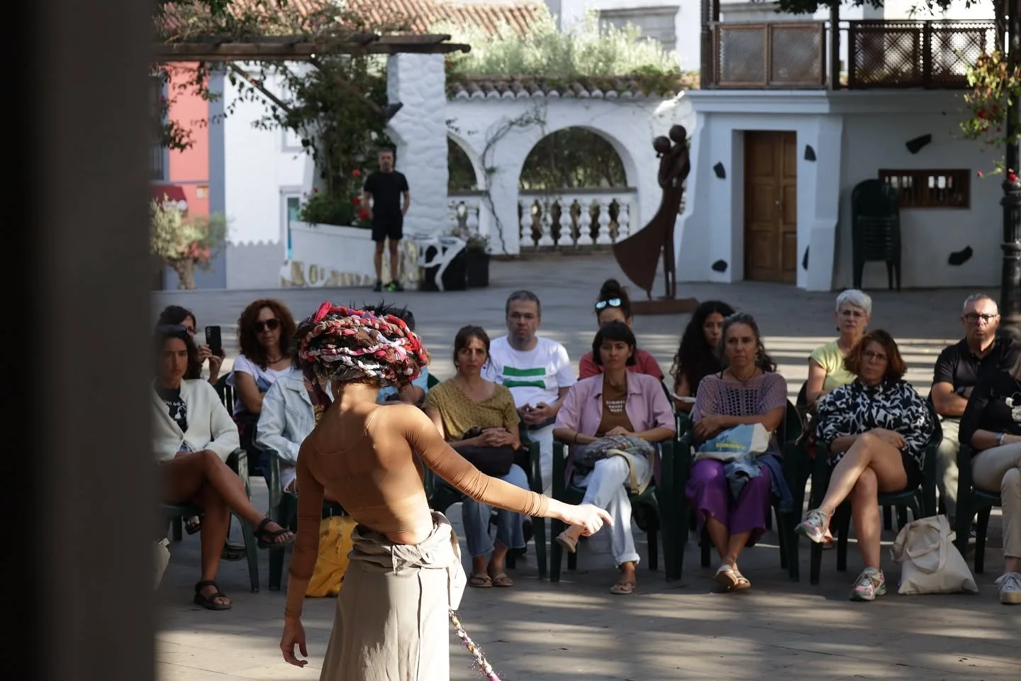 Una bailarina, de espaldas y con un tocado de tela de colores, actúa en una plaza ante un público sentado en semicírculo, con casas blancas y una escultura al fondo
