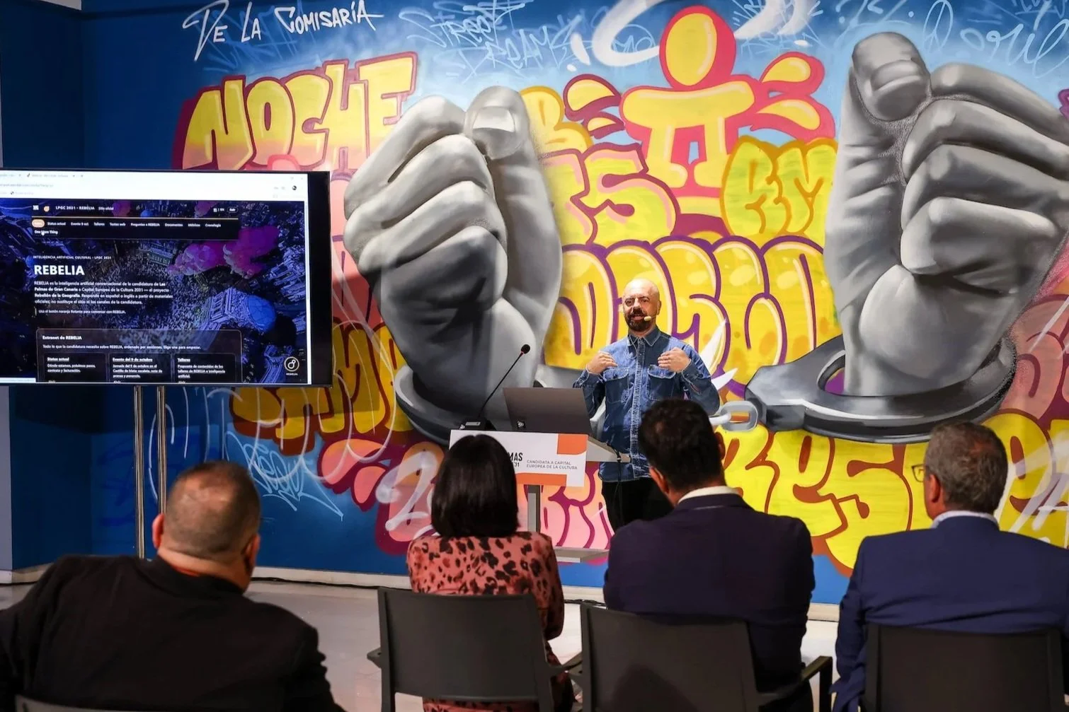 Pedro Mujica habla con las manos abiertas desde un atril, ante un mural de grafiti con puños de piedra y esposas y una pantalla que muestra la web de REBELIA, con el público de espaldas en primer plano
