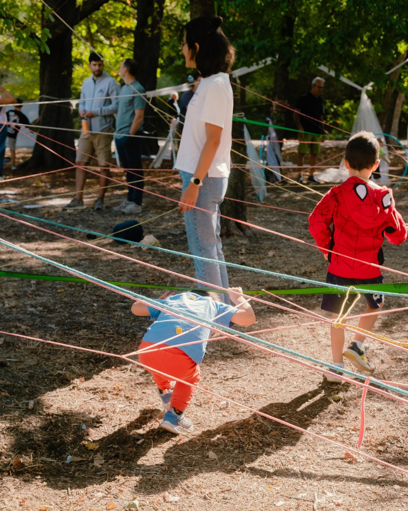Niños juegan agachándose y moviéndose entre una red de hilos de colores tensados entre los árboles de un parque, con adultos de pie al fondo