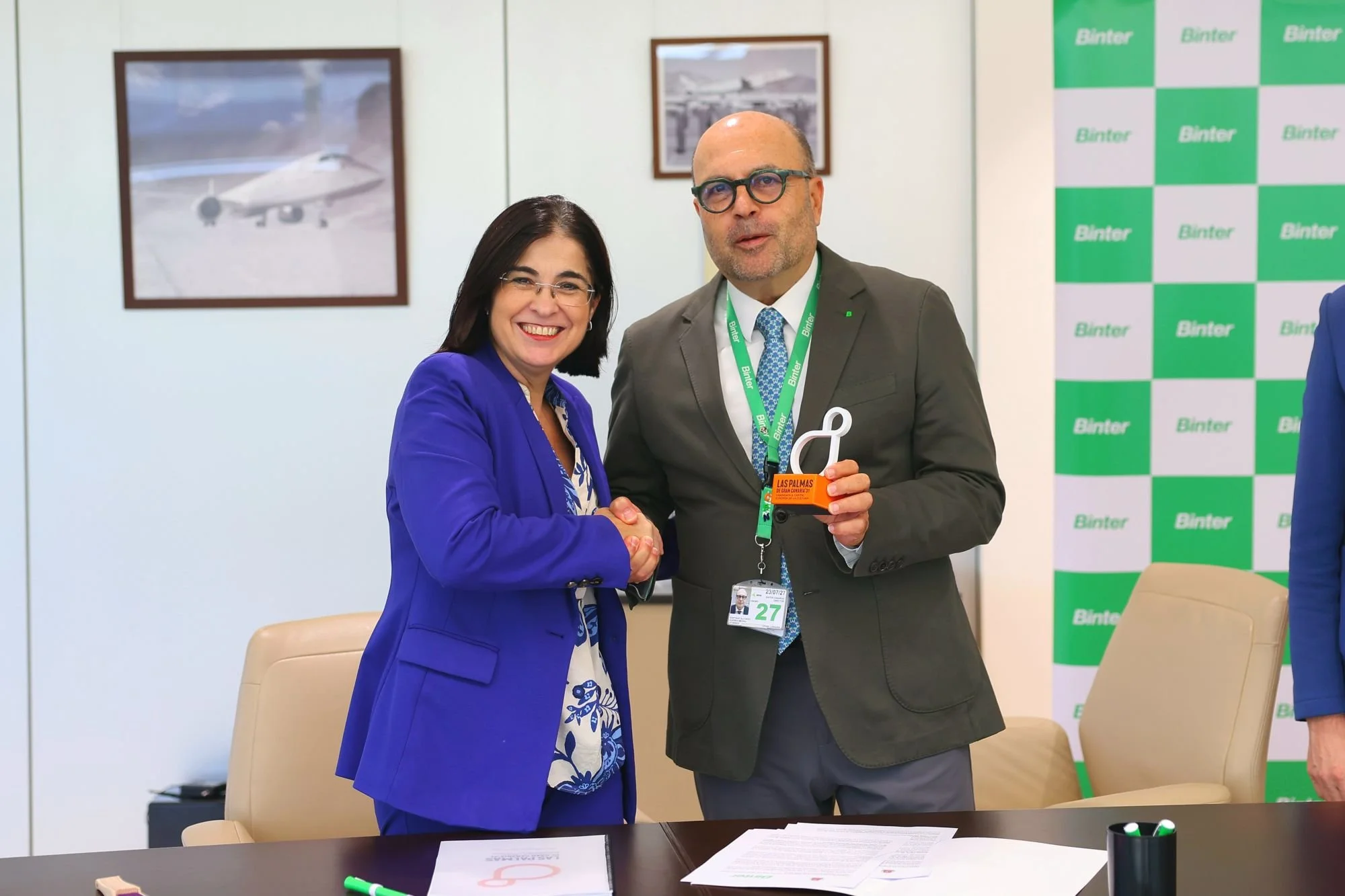 Carolina Darias estrecha la mano de Santiago Guerra, que sostiene la escultura del símbolo de LPGC’31, en un despacho con fotografías de aviones en la pared y un panel con el logotipo de Binter
