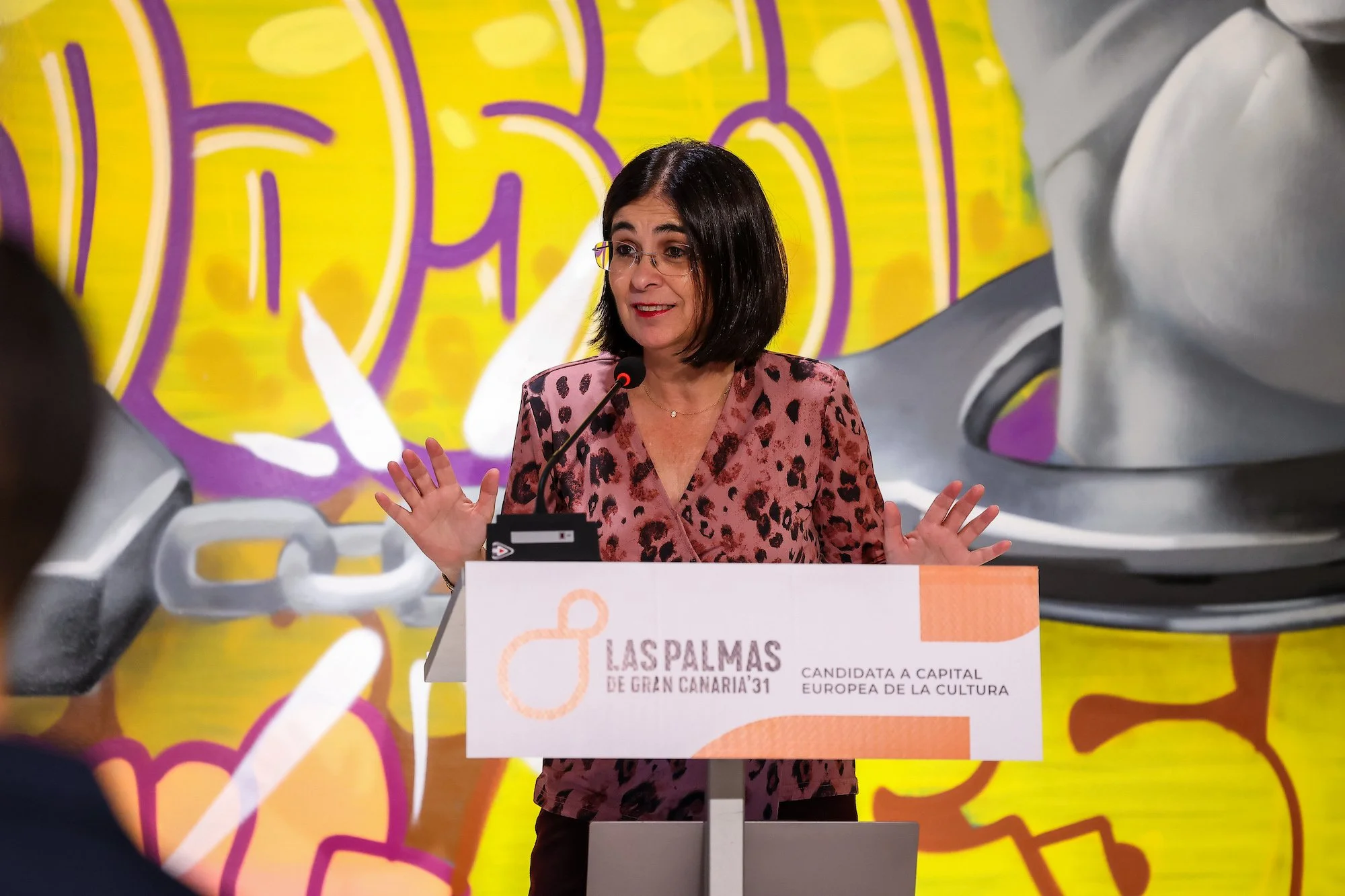 Carolina Darias gesticula con las manos abiertas tras un atril con el rótulo de la candidatura, ante un mural de grafiti de colores