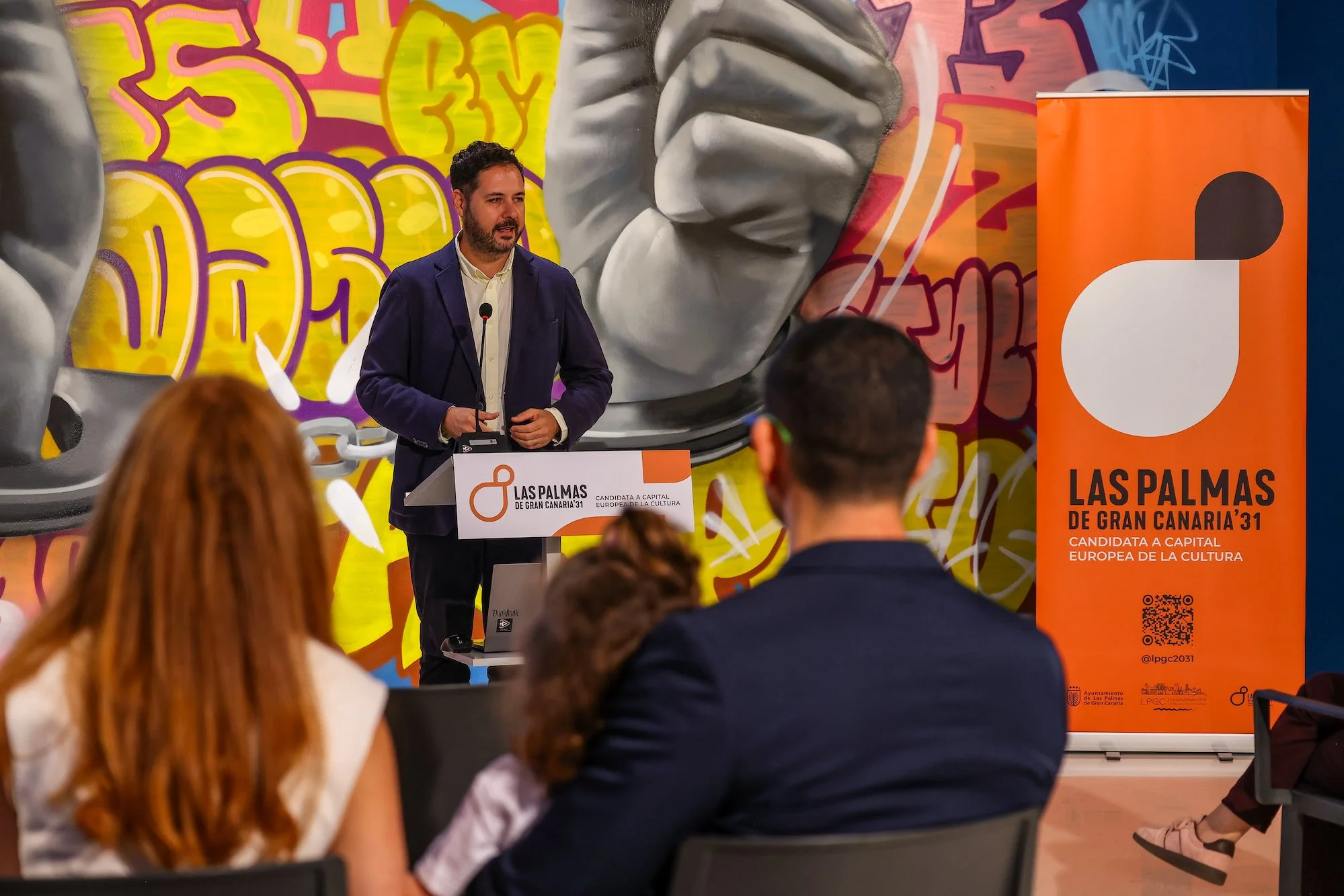 Josué Íñiguez habla desde un atril con el rótulo de la candidatura, ante un mural de grafiti, con un cartel naranja de LPGC’31 a la derecha y público de espaldas en primer plano