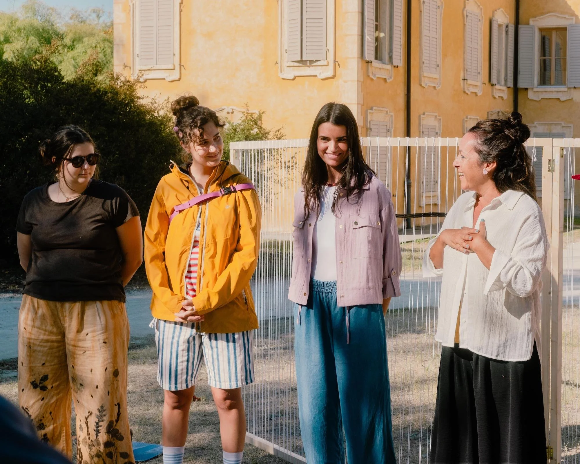 Cuatro mujeres conversan de pie al aire libre, frente a un edificio amarillo y una valla blanca con hilos verticales; la de la derecha habla sonriendo con las manos juntas