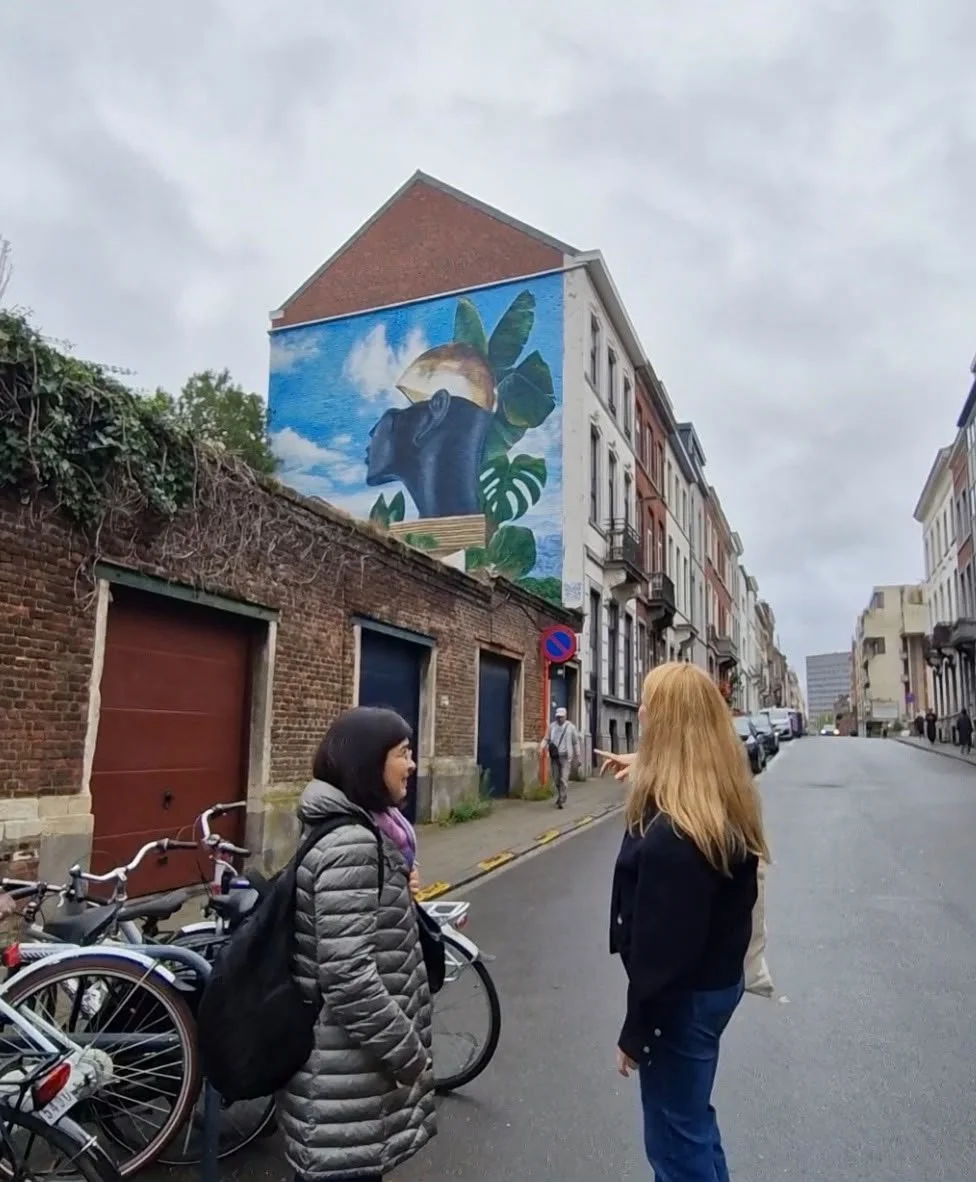 Dos mujeres, vistas de espaldas, miran en una calle de Leuven un gran mural pintado en el muro lateral de un edificio, con una figura de perfil y hojas grandes de monstera sobre un cielo azul; una de ellas lo señala con la mano
