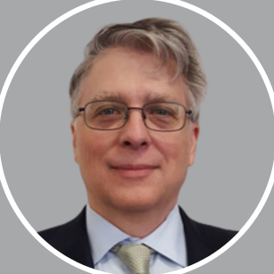 A middle-aged man with gray hair, wearing glasses, a dark suit, light blue shirt, and light-colored tie, smiling at the camera against a gray background.