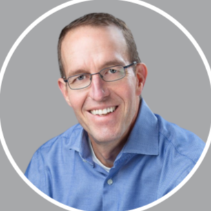 A man with glasses and short hair, smiling, wearing a blue button-up shirt, against a gray background.