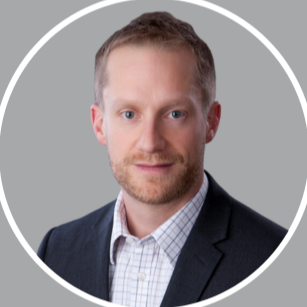 A professional headshot of a man with light brown hair, blue eyes, wearing a dark suit jacket and a light checkered shirt, against a gray background.