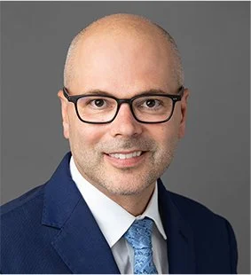 A professional headshot of a middle-aged man with glasses, wearing a dark blue suit, white dress shirt, and light blue tie, against a gray background.