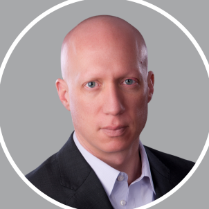 Headshot of a bald man in a black suit and white shirt against a gray background.