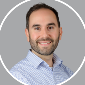 Portrait of a smiling man with dark hair and a beard, wearing a checkered shirt, in front of a gray background.