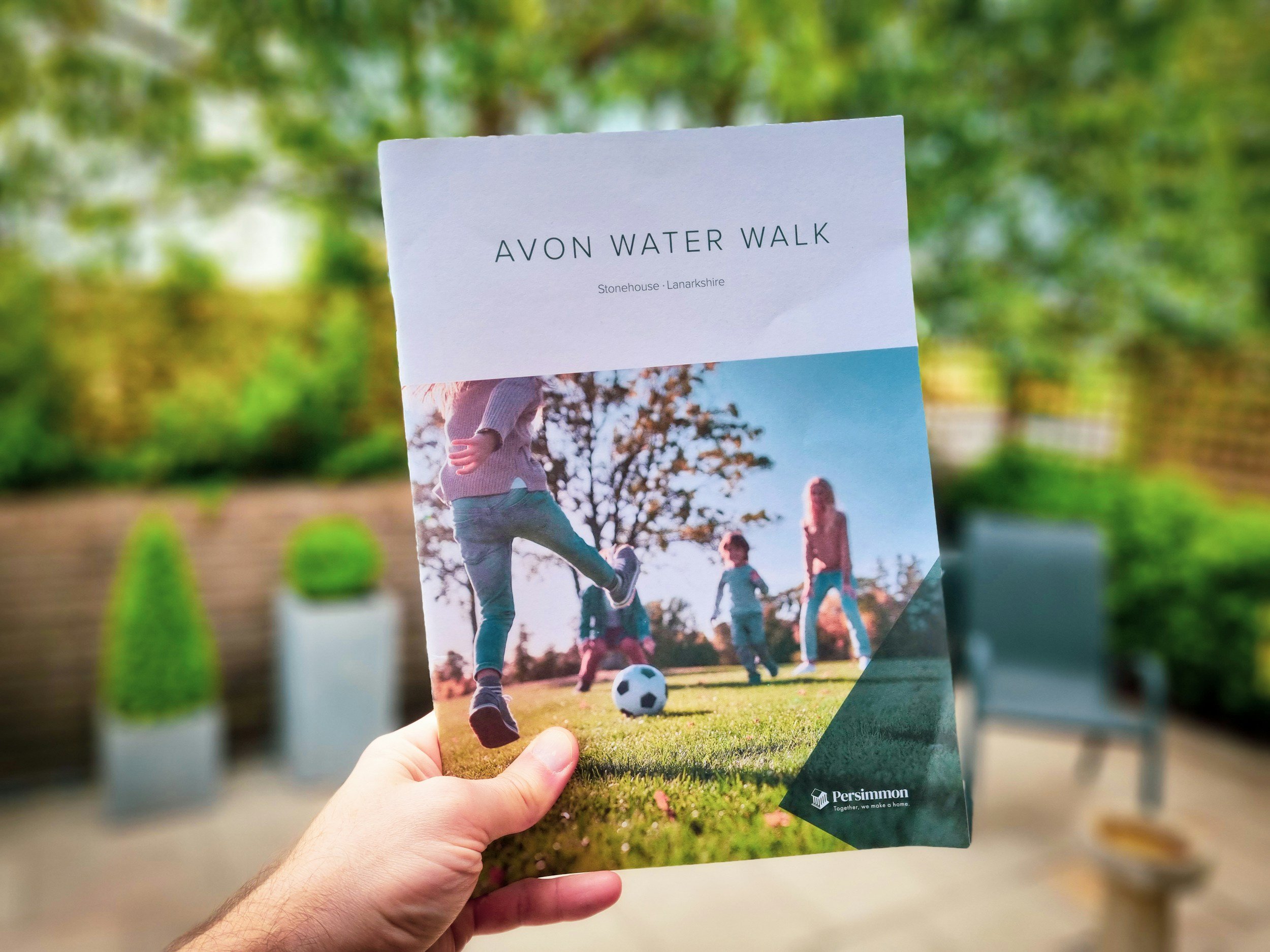 A person holding a brochure titled 'Avon Water Walk' with a picture of children playing soccer outdoors in a park with trees and greenery in the background.