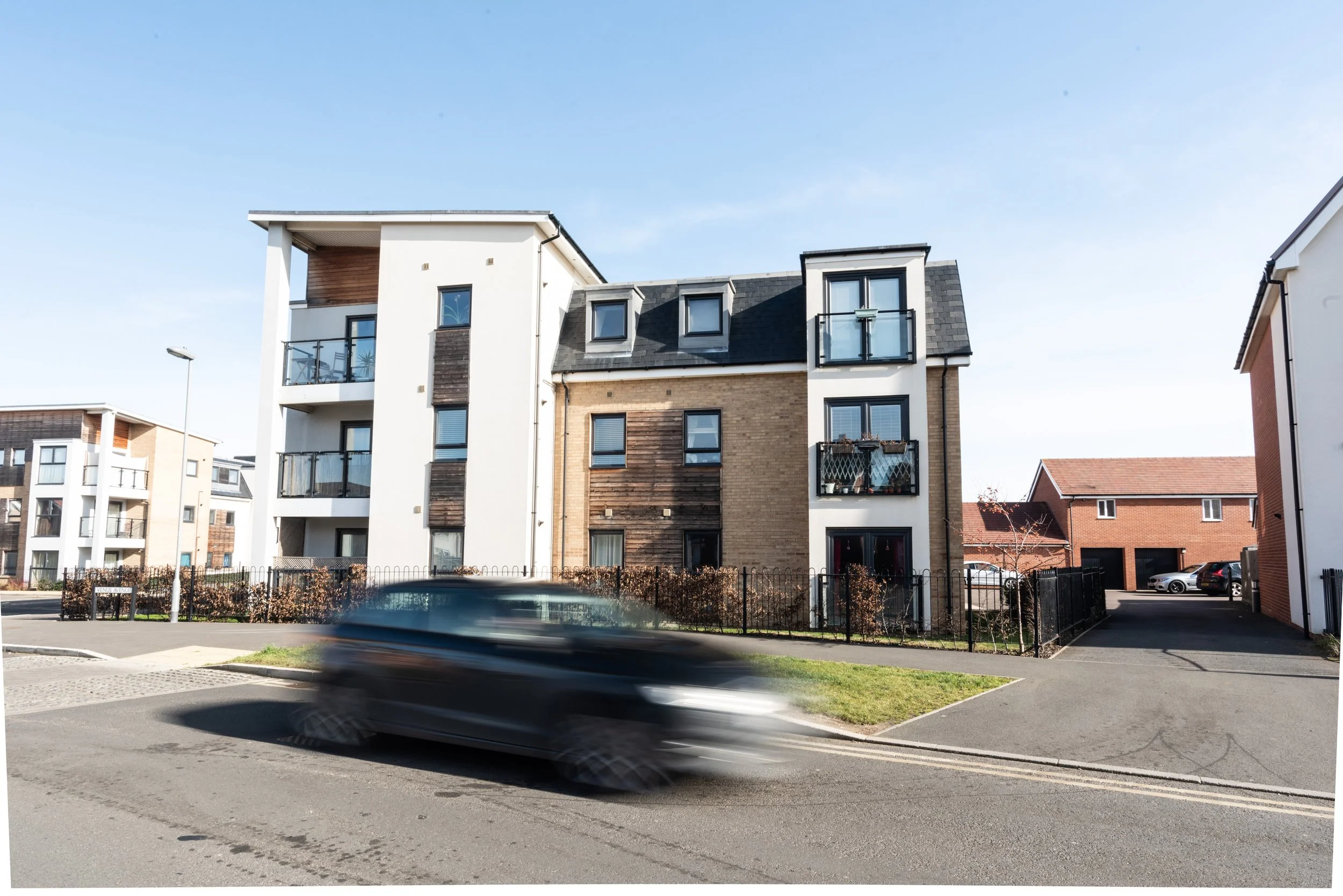 A modern apartment building with multiple floors, balconies, and various window styles. A car is passing in front of the building, appearing blurred due to motion. The building has mixed exterior materials, including wood and brick.