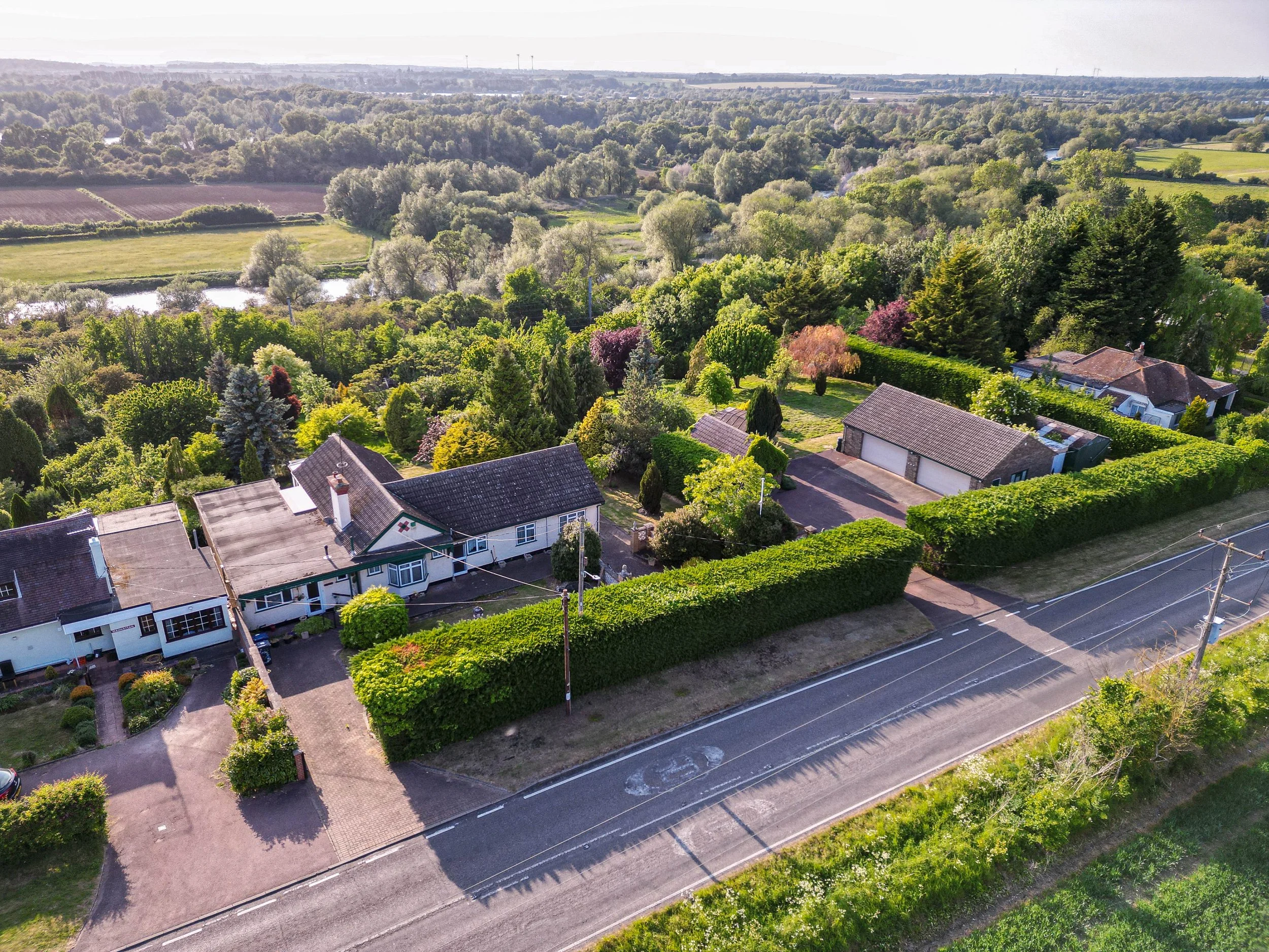 Aerial view of a suburban neighborhood with houses surrounded by lush green trees and gardens, next to a river and open fields under a clear sky.