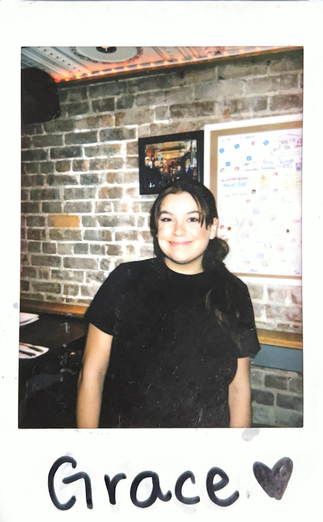 Person smiling in front of a brick wall background with framed artwork and a map on the wall.