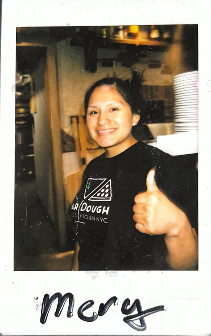 Person smiling and giving a thumbs-up, wearing a black "SOURDOUGH PIZZA KITCHEN NYC" t-shirt, standing indoors.