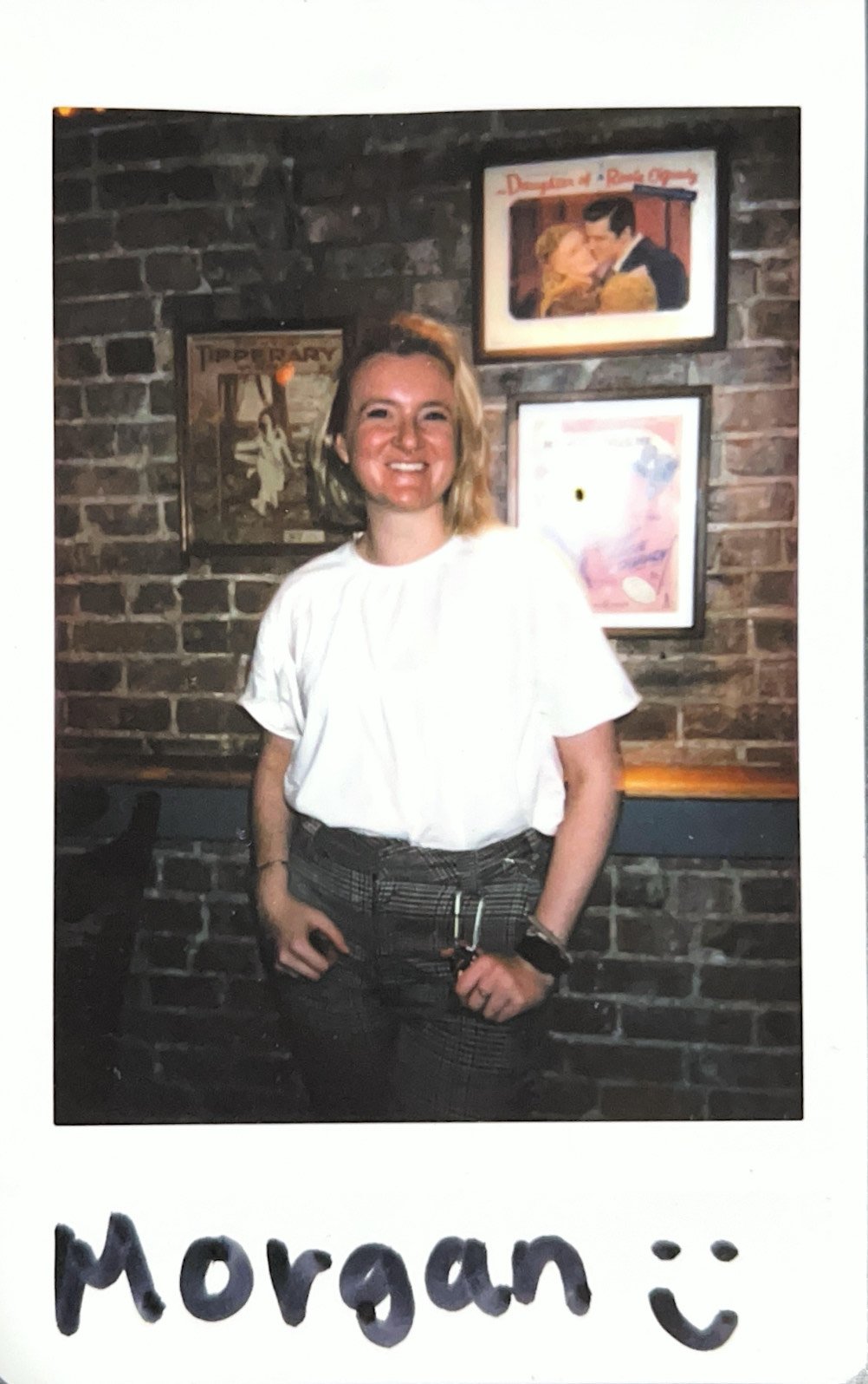 Person standing in front of a brick wall with framed vintage posters, wearing a white shirt and plaid pants, smiling at the camera. Handwritten note "Morgan :)" at the bottom.