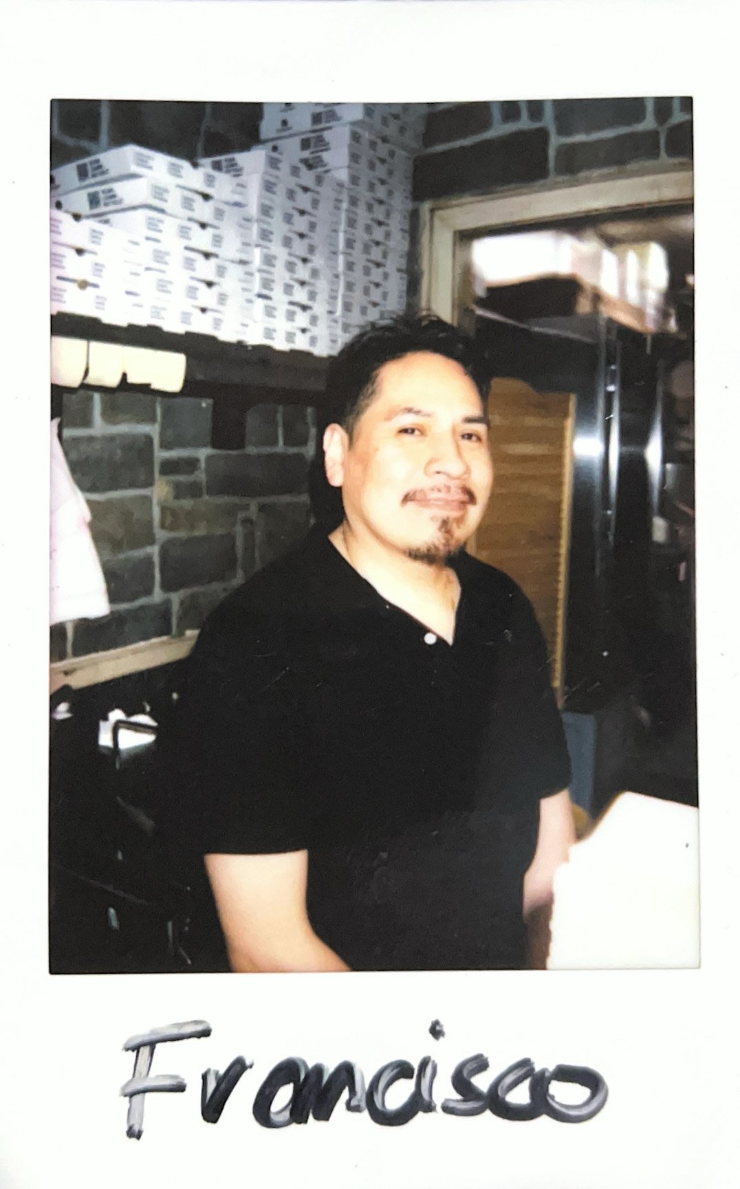 Person in black shirt smiling in a room with brick walls and pizza boxes.