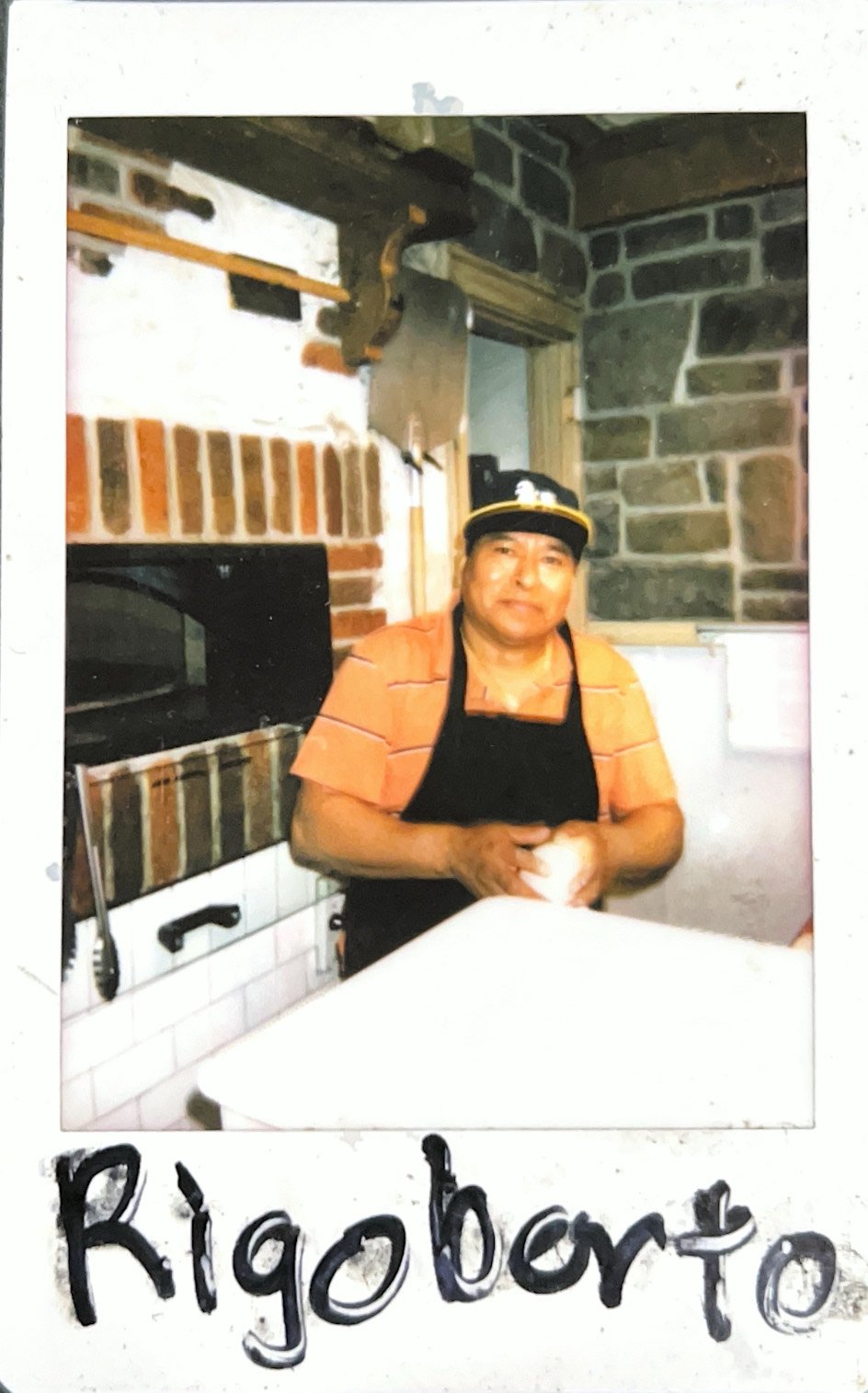 Person wearing a hat and apron standing in a kitchen with a brick oven, holding a ball of dough. Text below reads "Rigoberto."