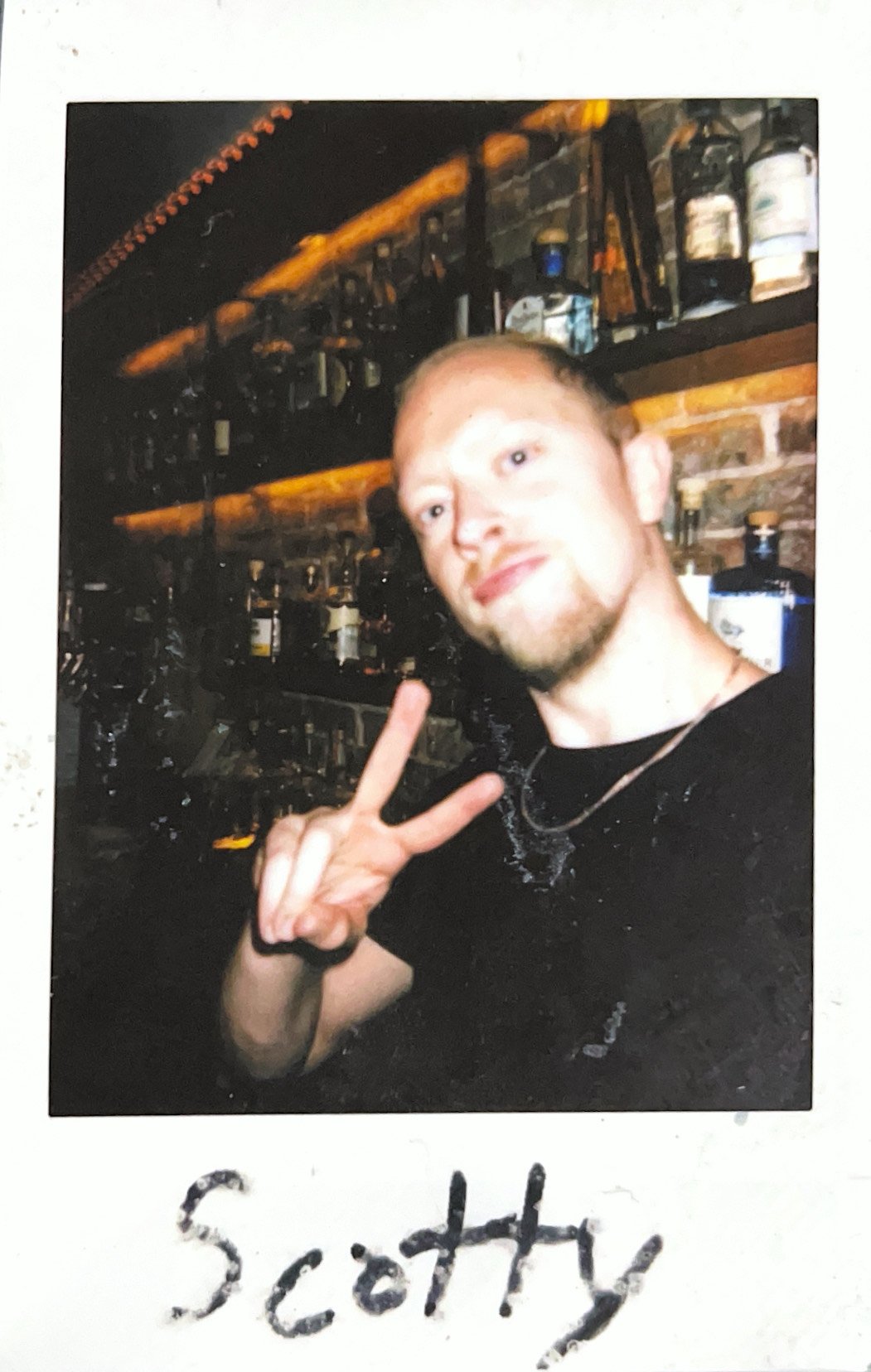 Person in a bar making a peace sign with bottles on shelves in the background.