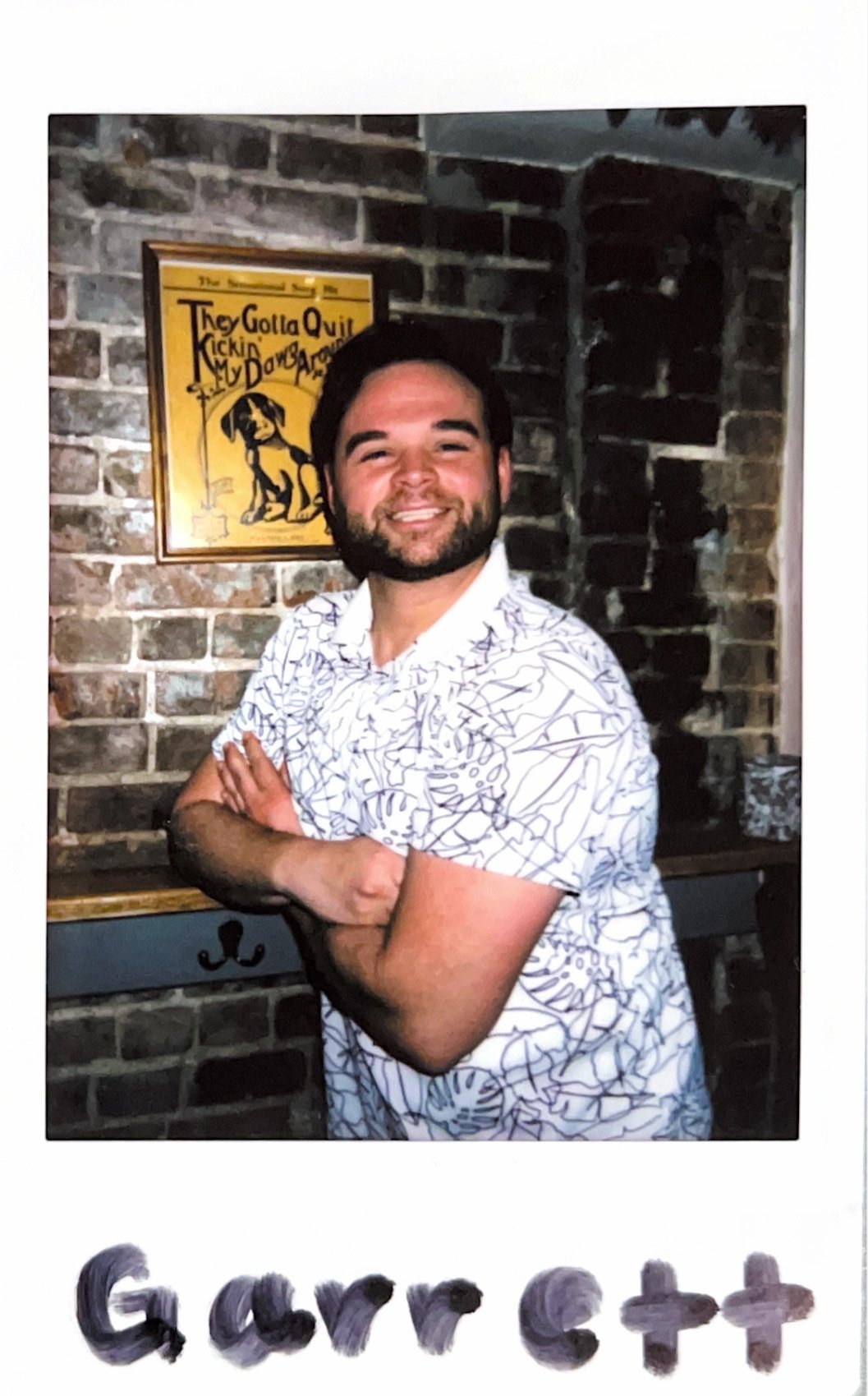 Smiling person in a patterned shirt posing in front of a brick wall with a vintage sign in the background.